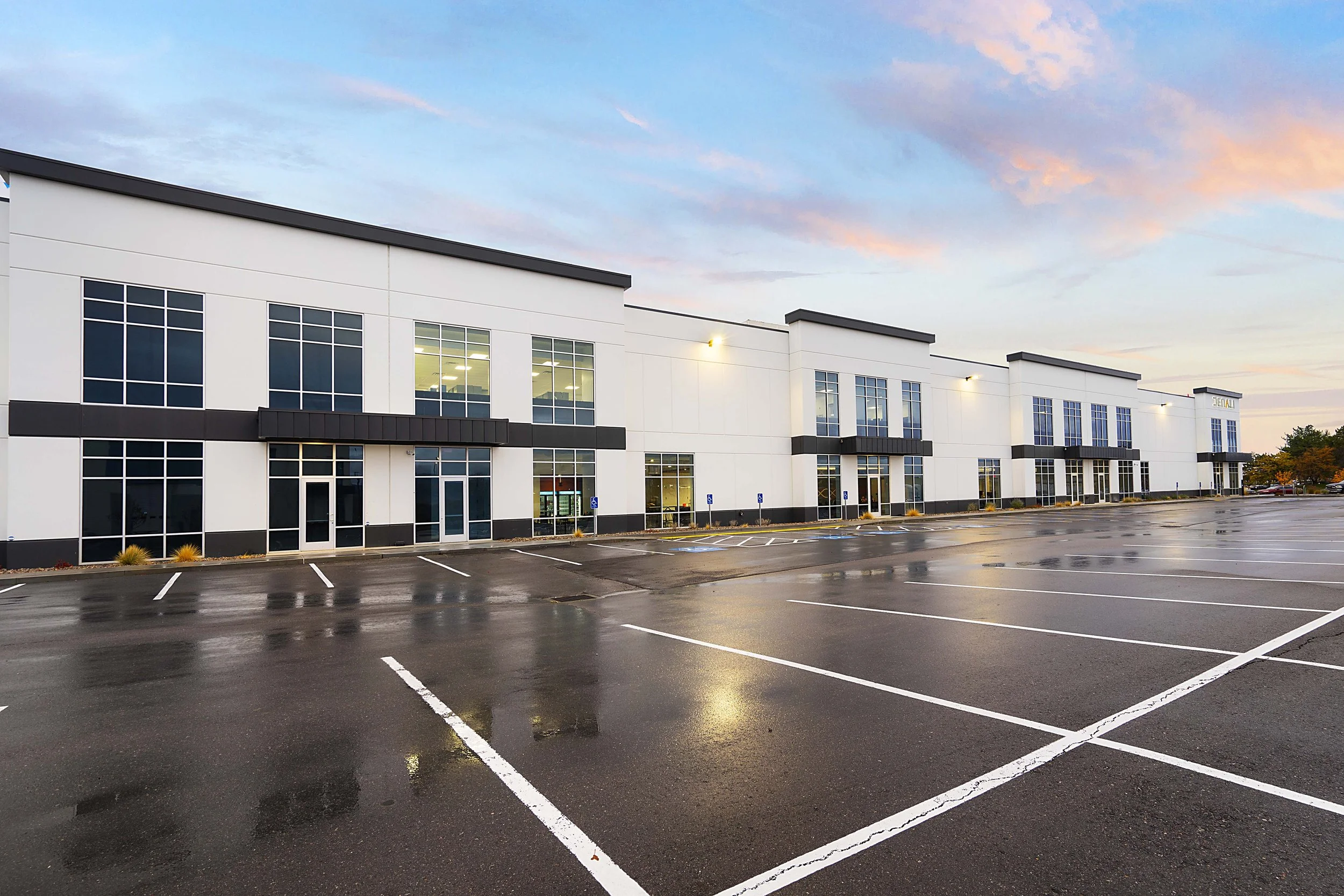 Empty parking lot outside a modern, white commercial building with large windows, during dusk or early evening, with some clouds and wet pavement reflecting the building lights.