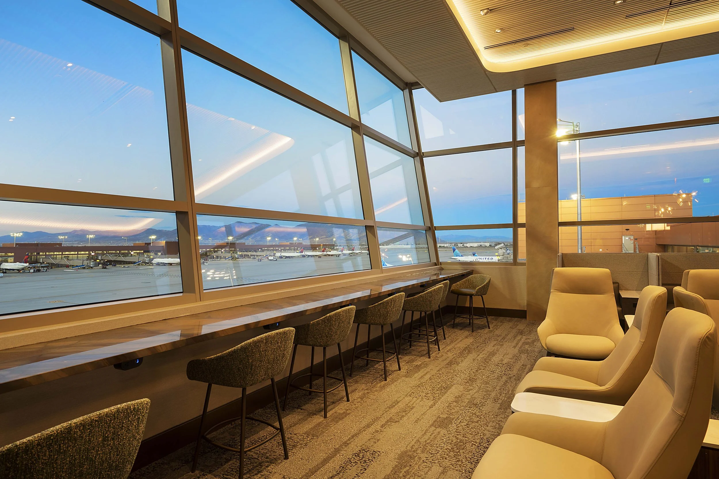 Airport lounge with large windows overlooking airplanes on the tarmac during sunset, featuring beige chairs and a long counter with seats.