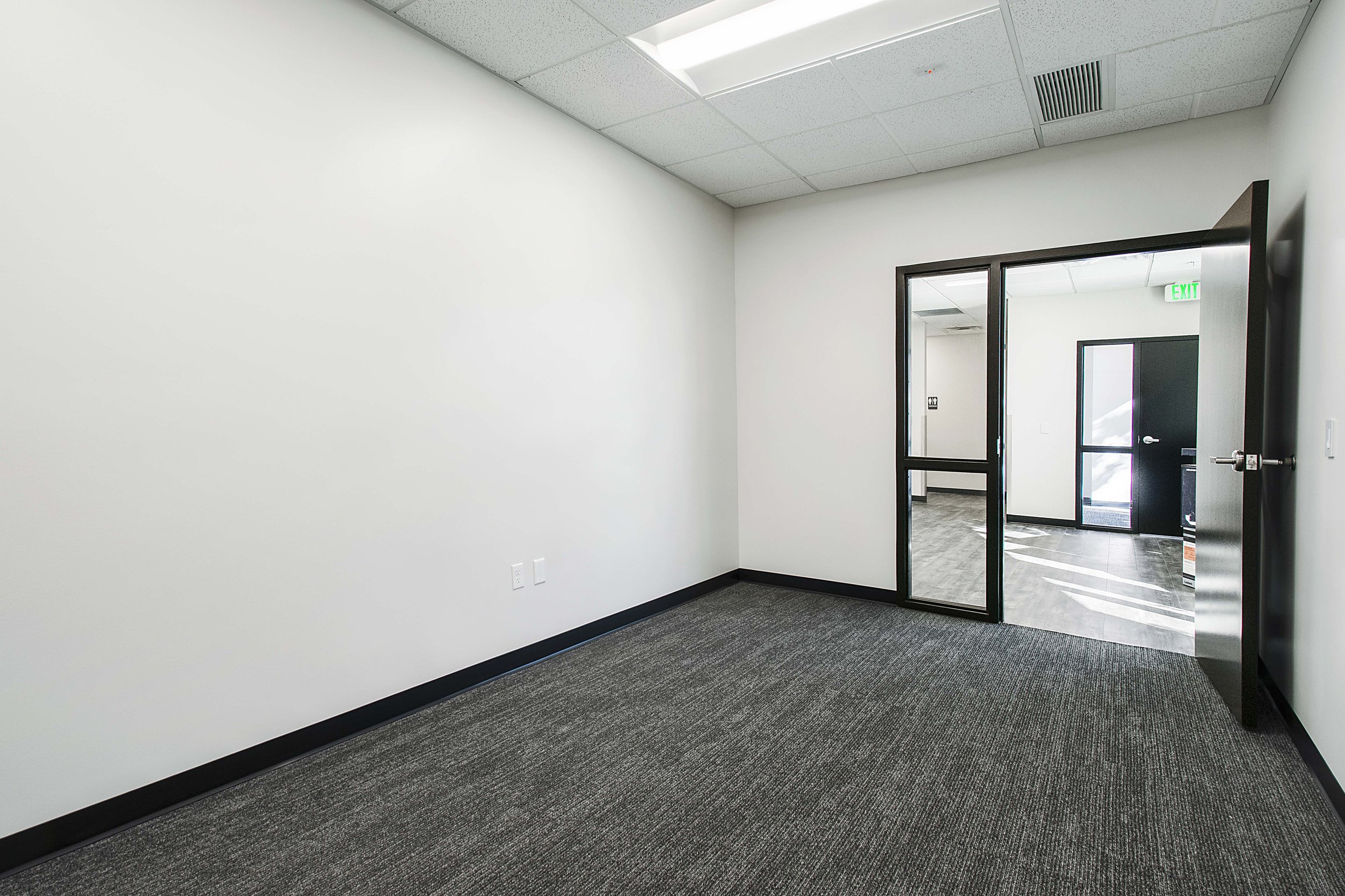Empty office room with white walls, dark carpet, double glass doors, and exit sign in hallway.