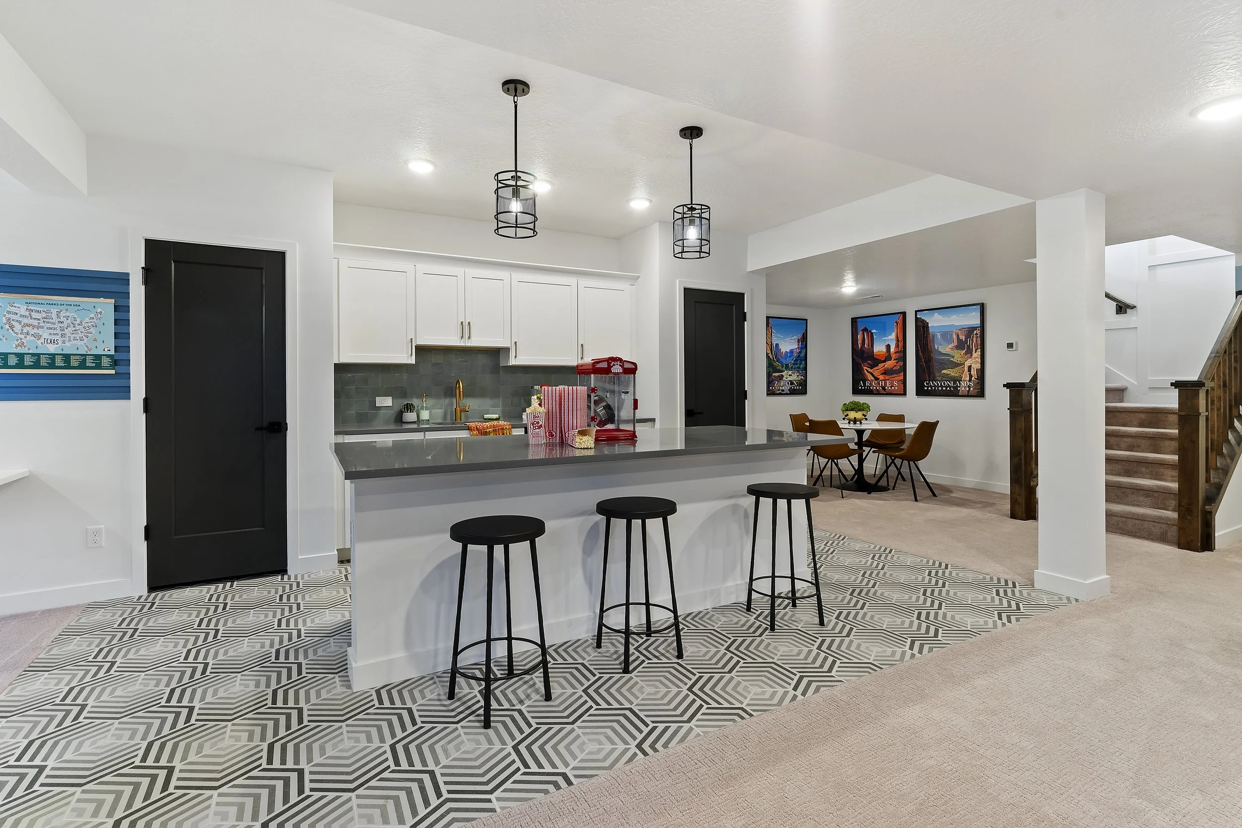 Open-concept kitchen with white cabinets, gray countertops, black door, patterned tile floor, three black barstools, and a small dining area with three brown chairs and framed landscape art on the wall.