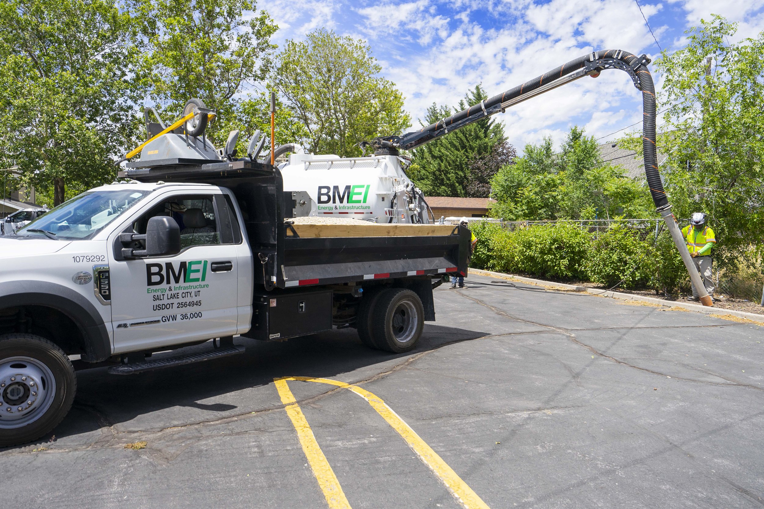 A utility truck from BMEI Energy & Infrastructure is parked in a lot, with a worker wearing a helmet and safety vest fixing a fallen utility pole or tree branch.