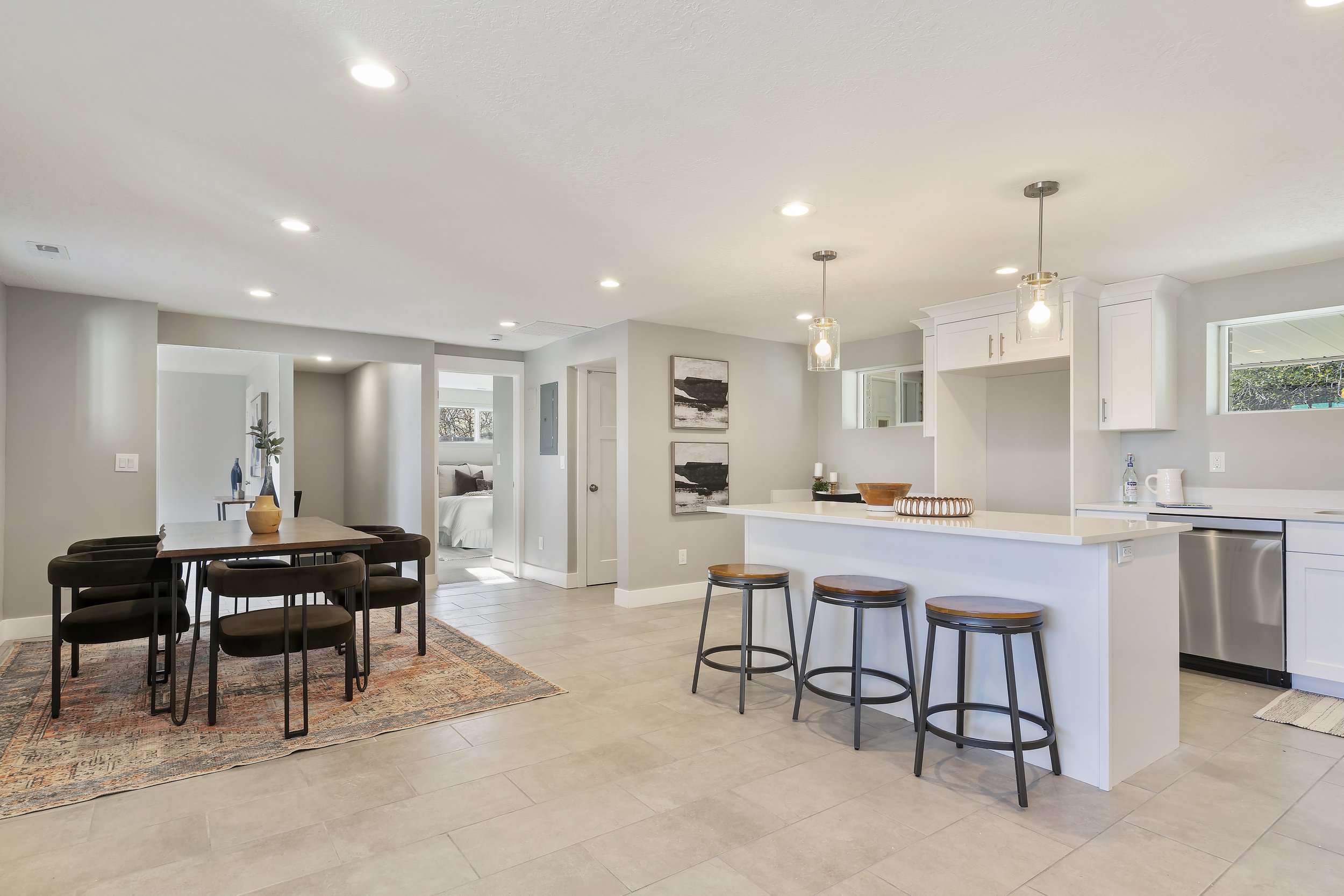 Open-concept kitchen and dining area with white cabinets, black bar stools at a white kitchen island, a wooden dining table with black chairs, and artwork on light gray walls.