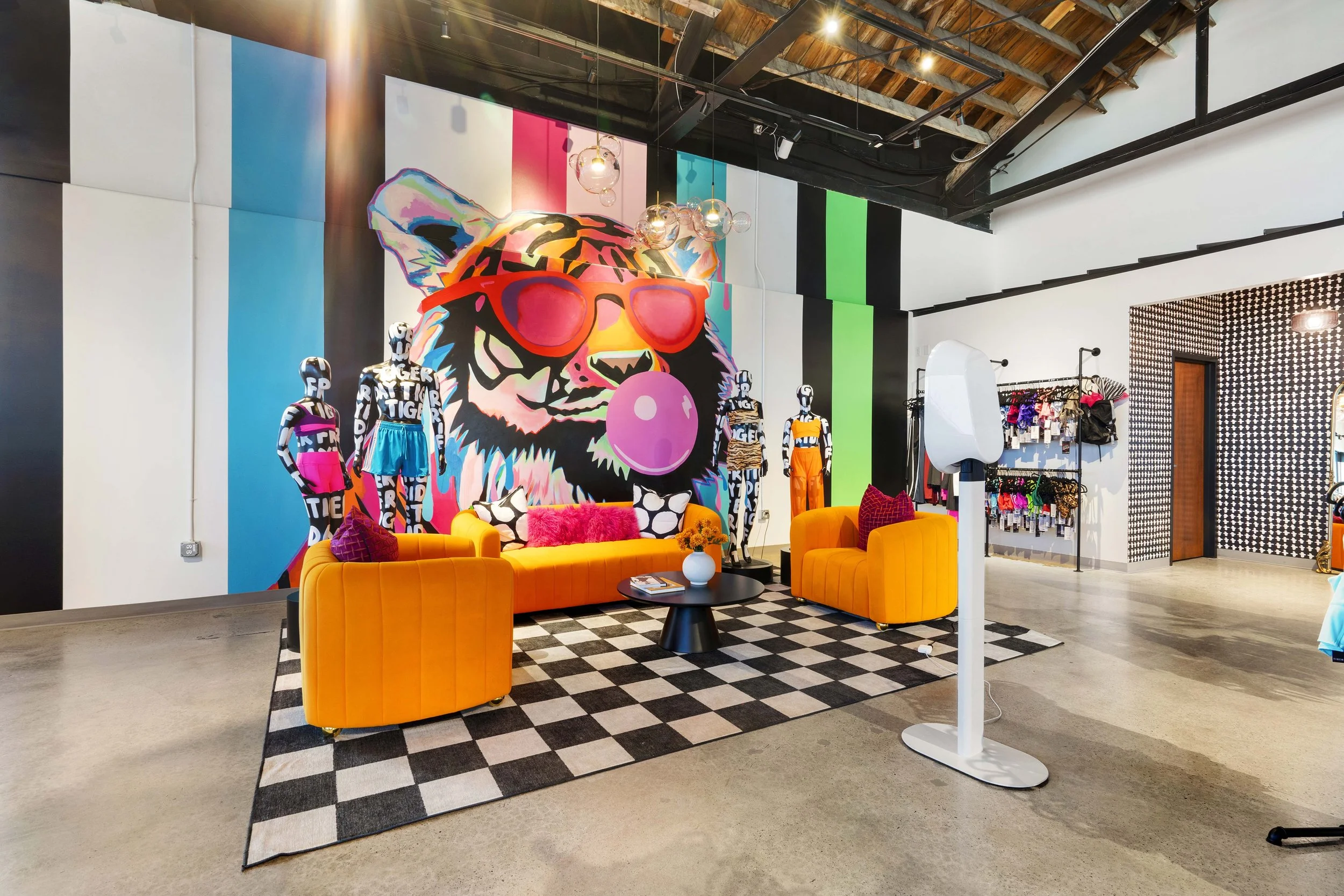 Colorful boutique interior with a large mural of a tiger wearing sunglasses and blowing a bubble gum, surrounded by mannequins dressed in vibrant clothes, orange sofas, a black and white checkered rug, and various display racks.
