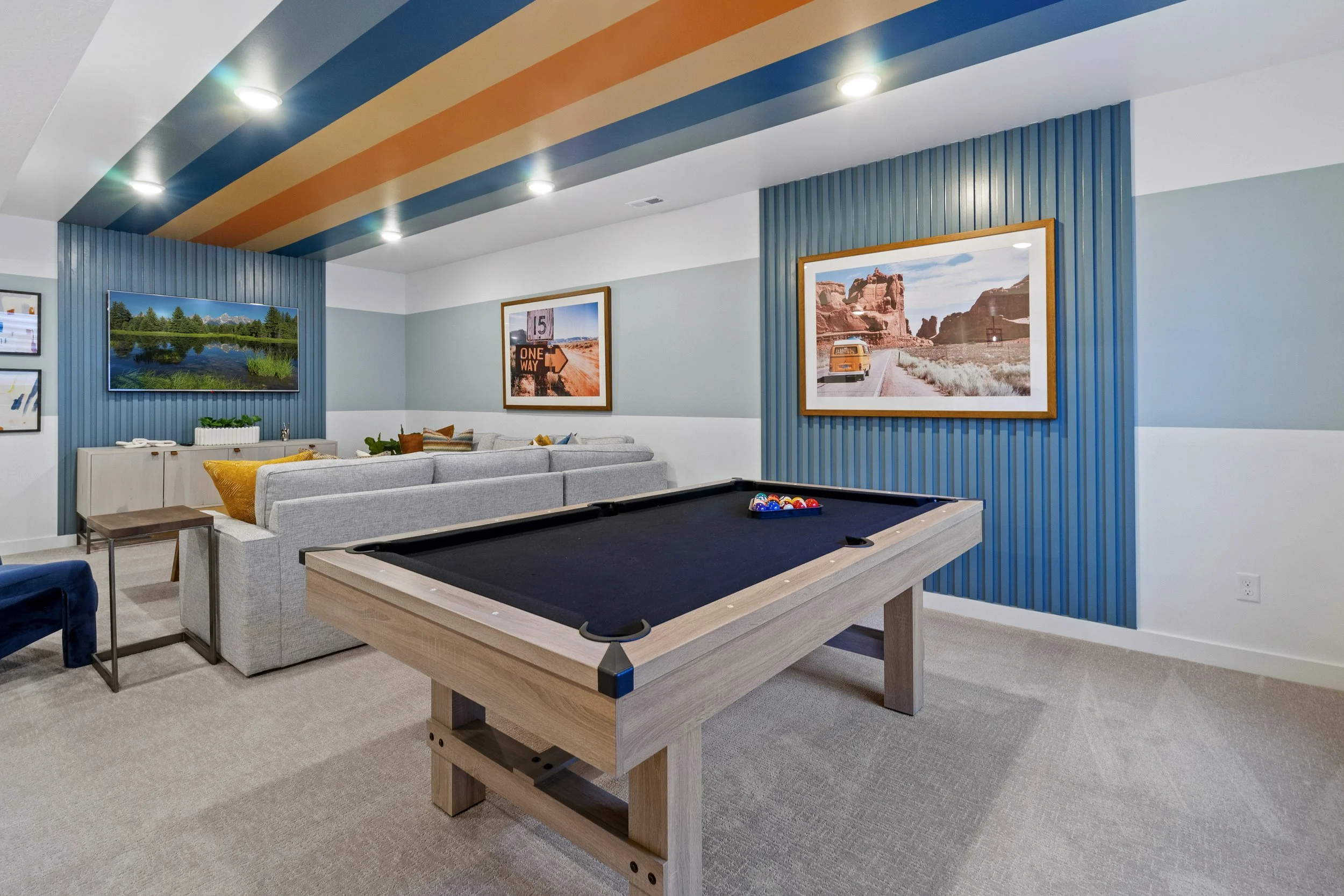 A game room with a pool table in the foreground, a gray sofa with yellow and patterned pillows behind it, framed pictures on the walls, and ceiling lights. The ceiling has colorful stripes.