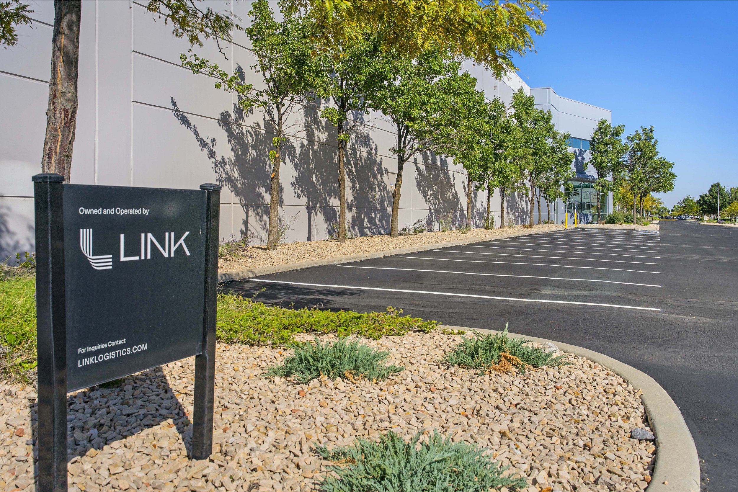 Empty parking lot outside a modern building with trees and a sign that reads 'Owned and Operated by LINK' and a contact website LINKLOGISTICS.COM.