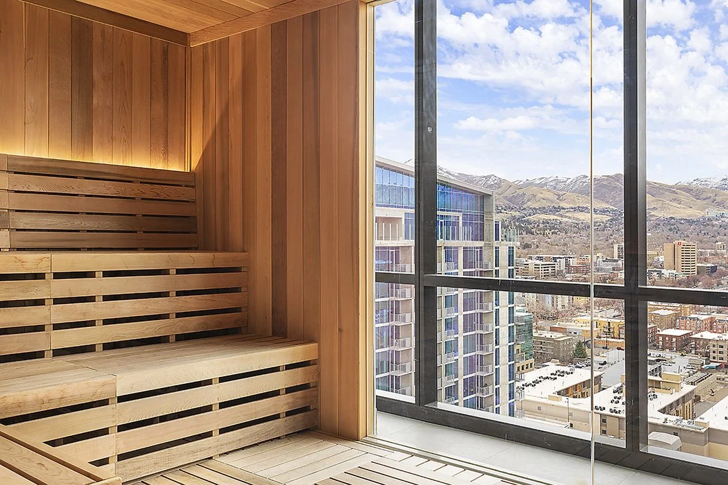 Interior of a sauna with wooden benches, next to a large window showing a cityscape and mountains in the background.