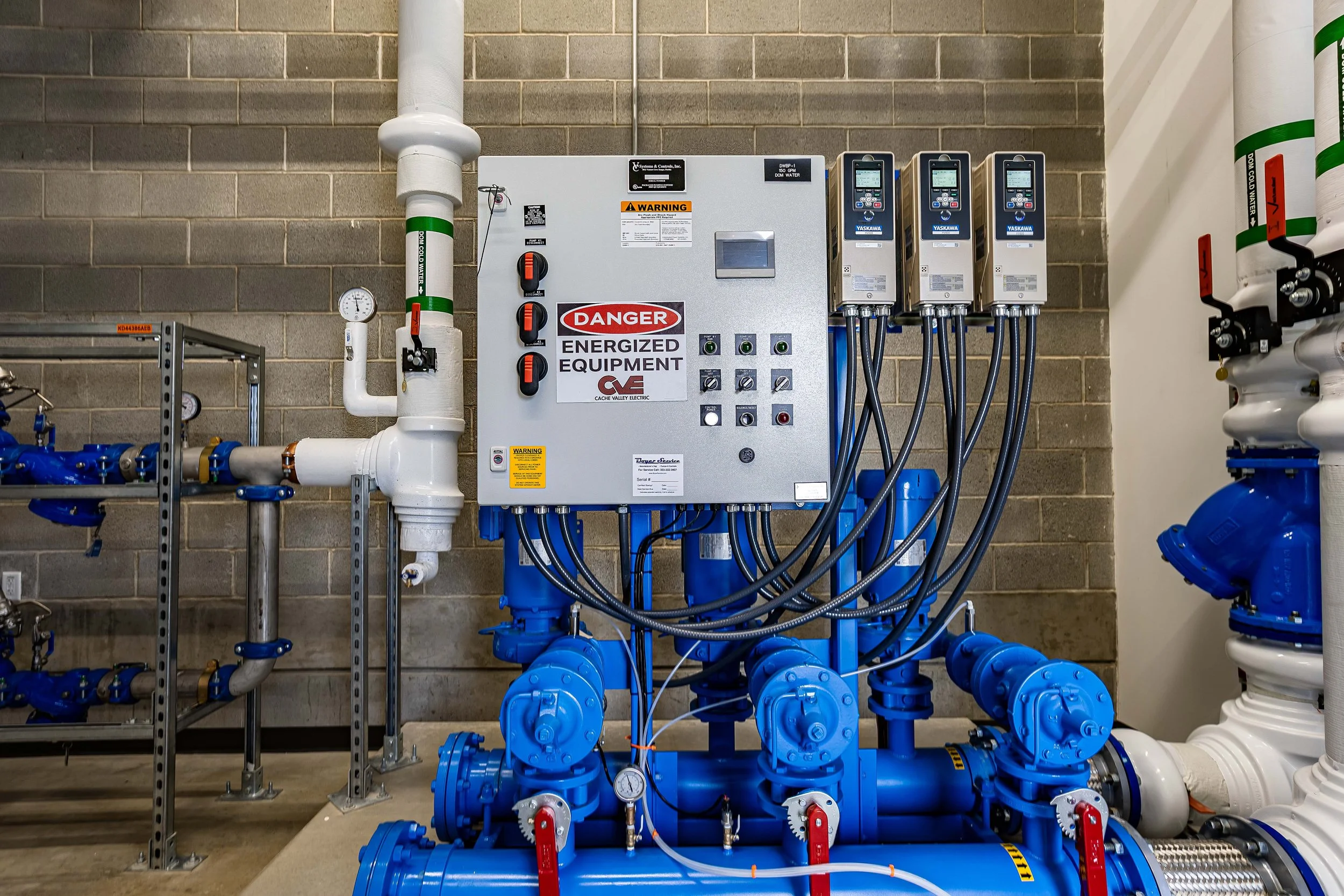 An industrial utility room containing a large electrical control panel labeled "Danger Energized Equipment" with warning signs, gauges, and several connected blue pipes and pumps.