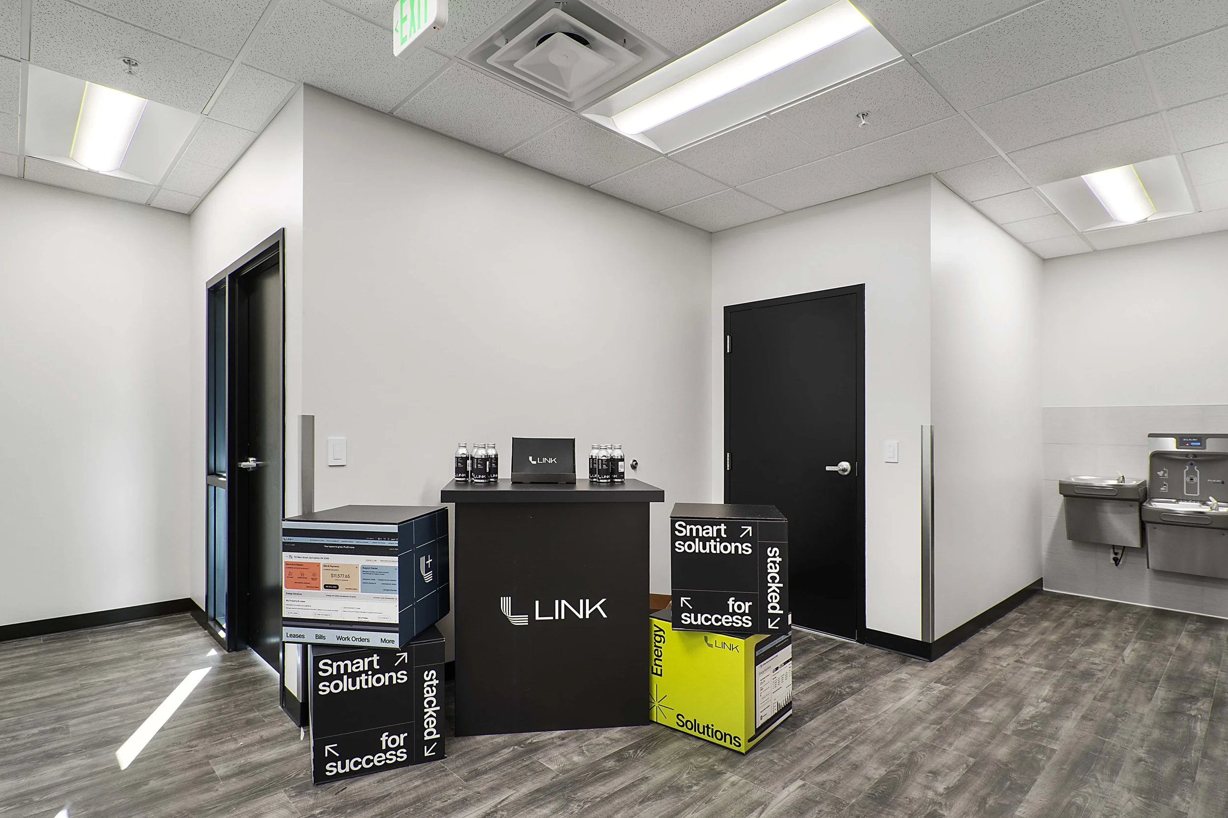 Reception area with a black desk featuring the 'LINK' logo, water bottles on top, promotional boxes with phrases like 'Smart solutions' and 'Energy Solutions', and a hallway with water fountains on the right. The ceiling has fluorescent lights and an