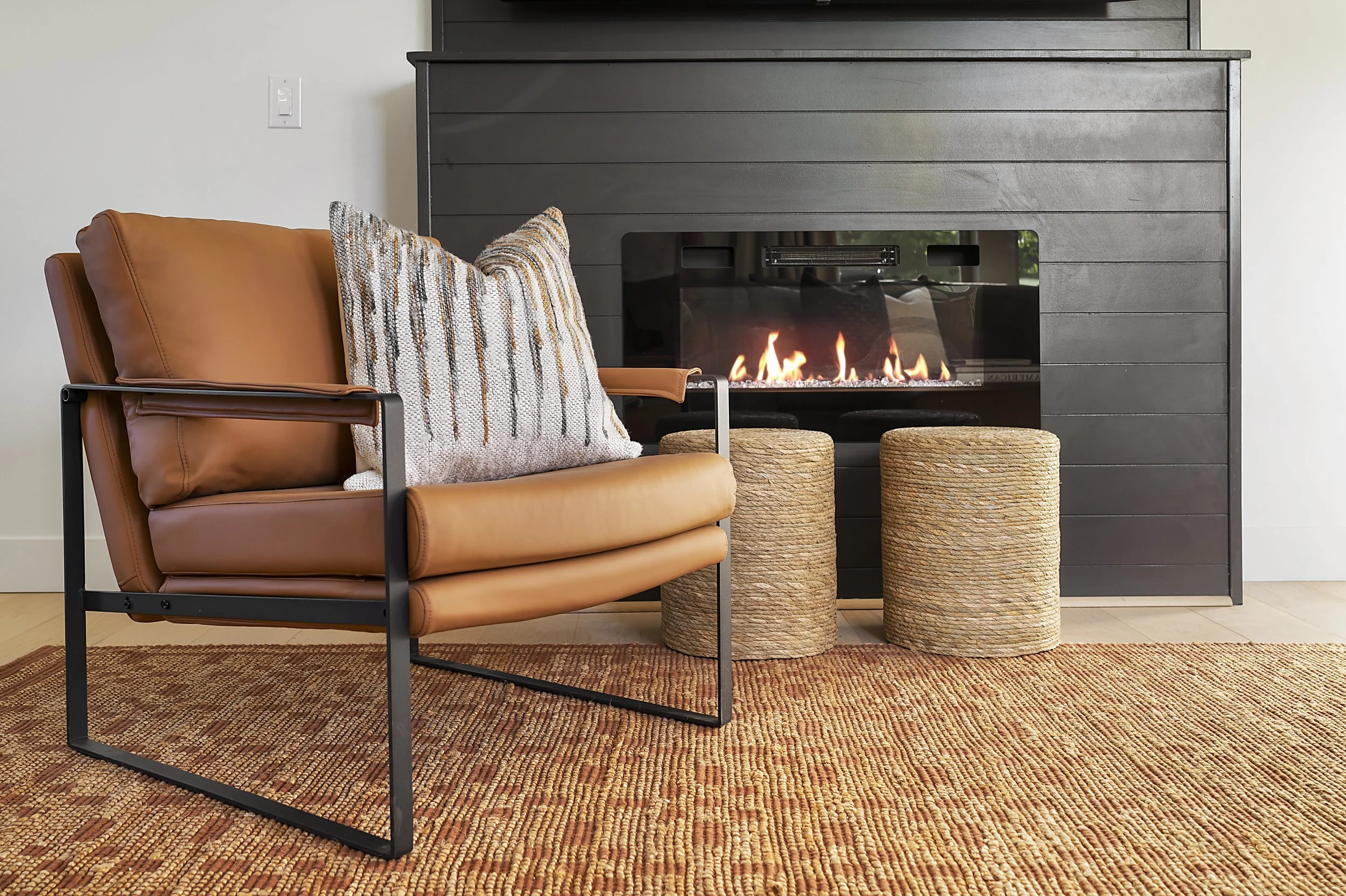 Modern living room with a tan leather armchair, a decorative pillow, a black fireplace with a lit fire, two woven ottomans, a rust-colored rug, and a white wall.