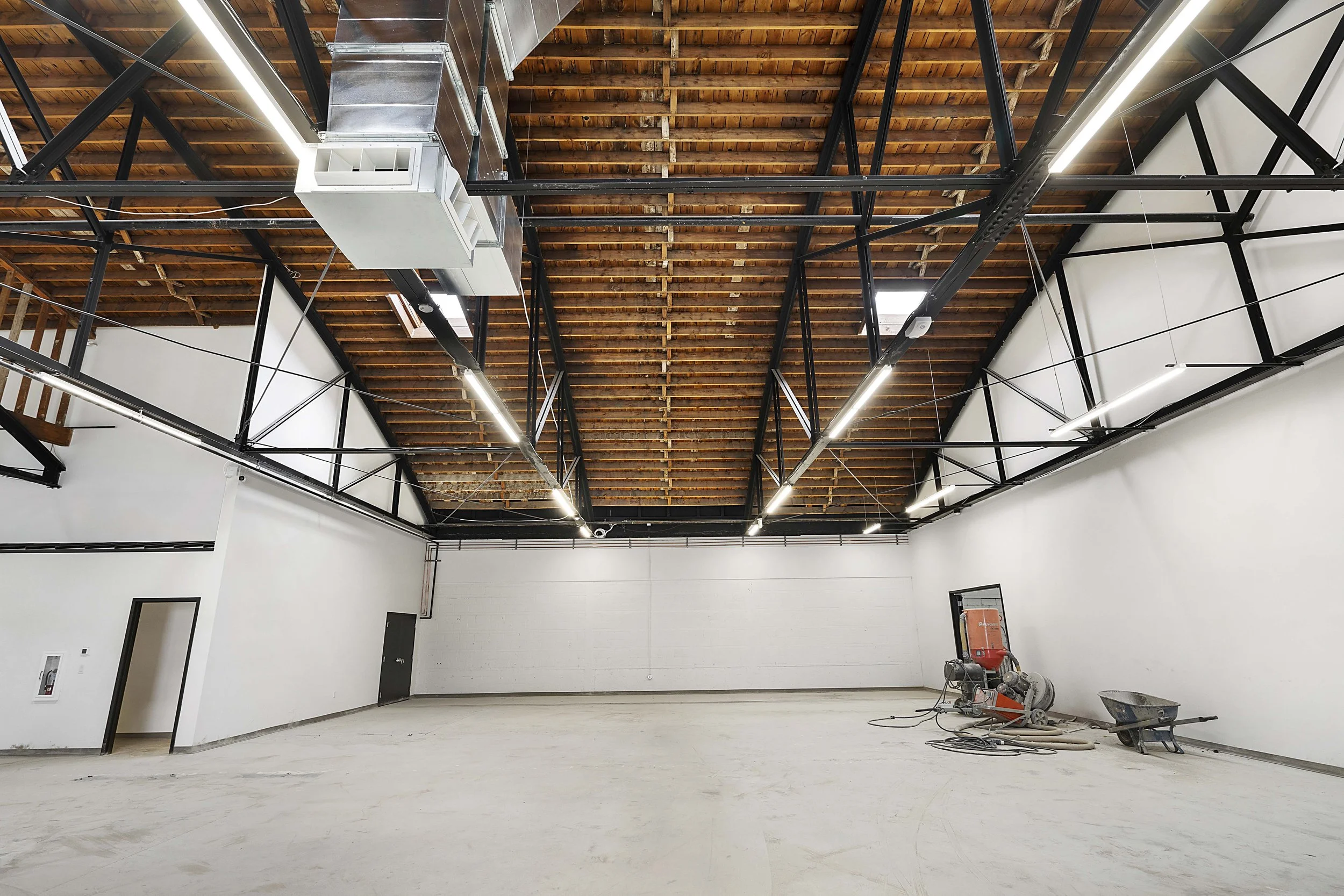 Large interior space under construction with white walls, unfinished flooring, black metal ceiling framework, and exposed wooden ceiling. There are construction tools and equipment on the floor, including a wheelbarrow, a cement mixer, and cables.