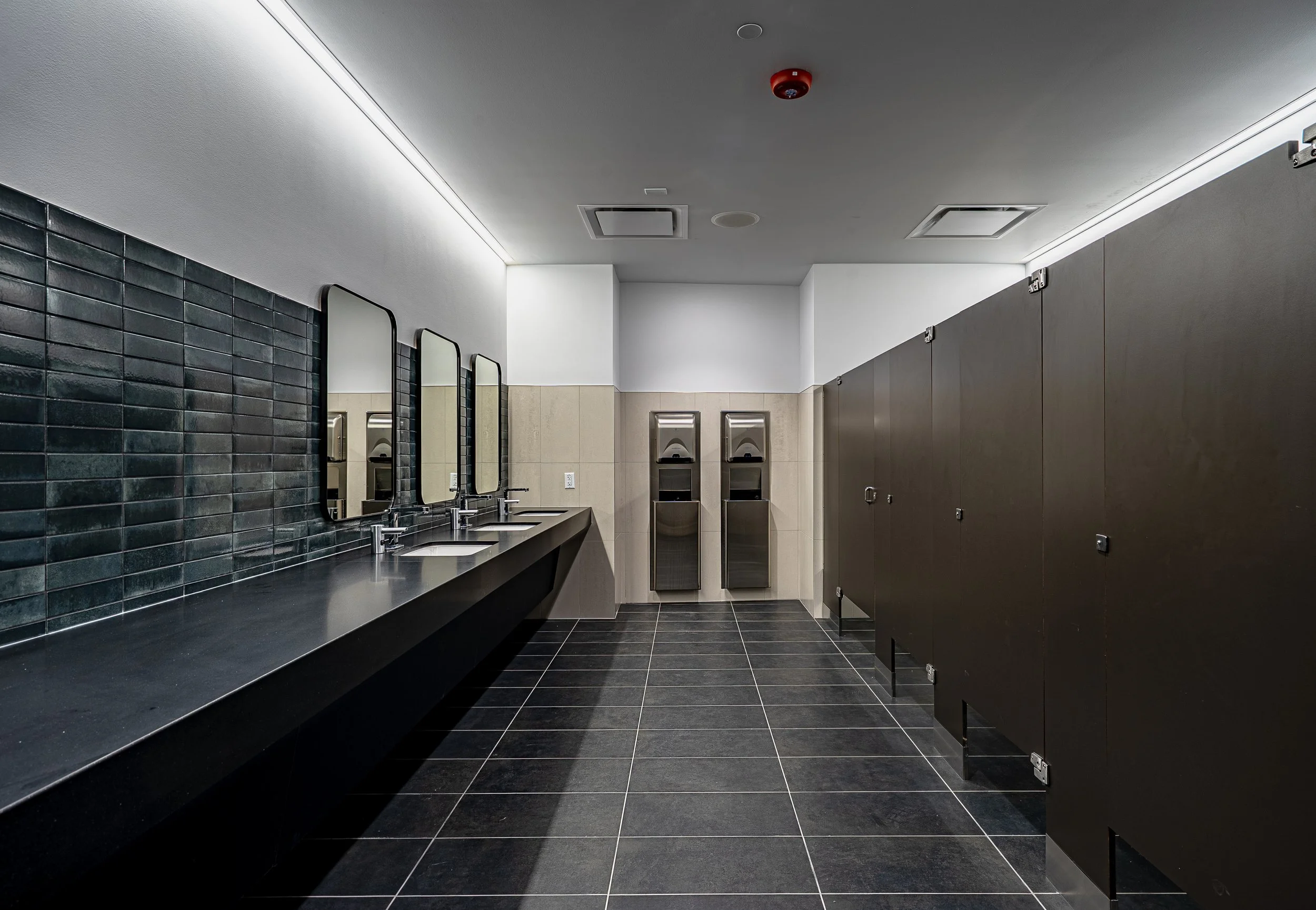 A modern public restroom with four sinks, three mirrors, and three hand dryers, with black tiled floors and black stalls.