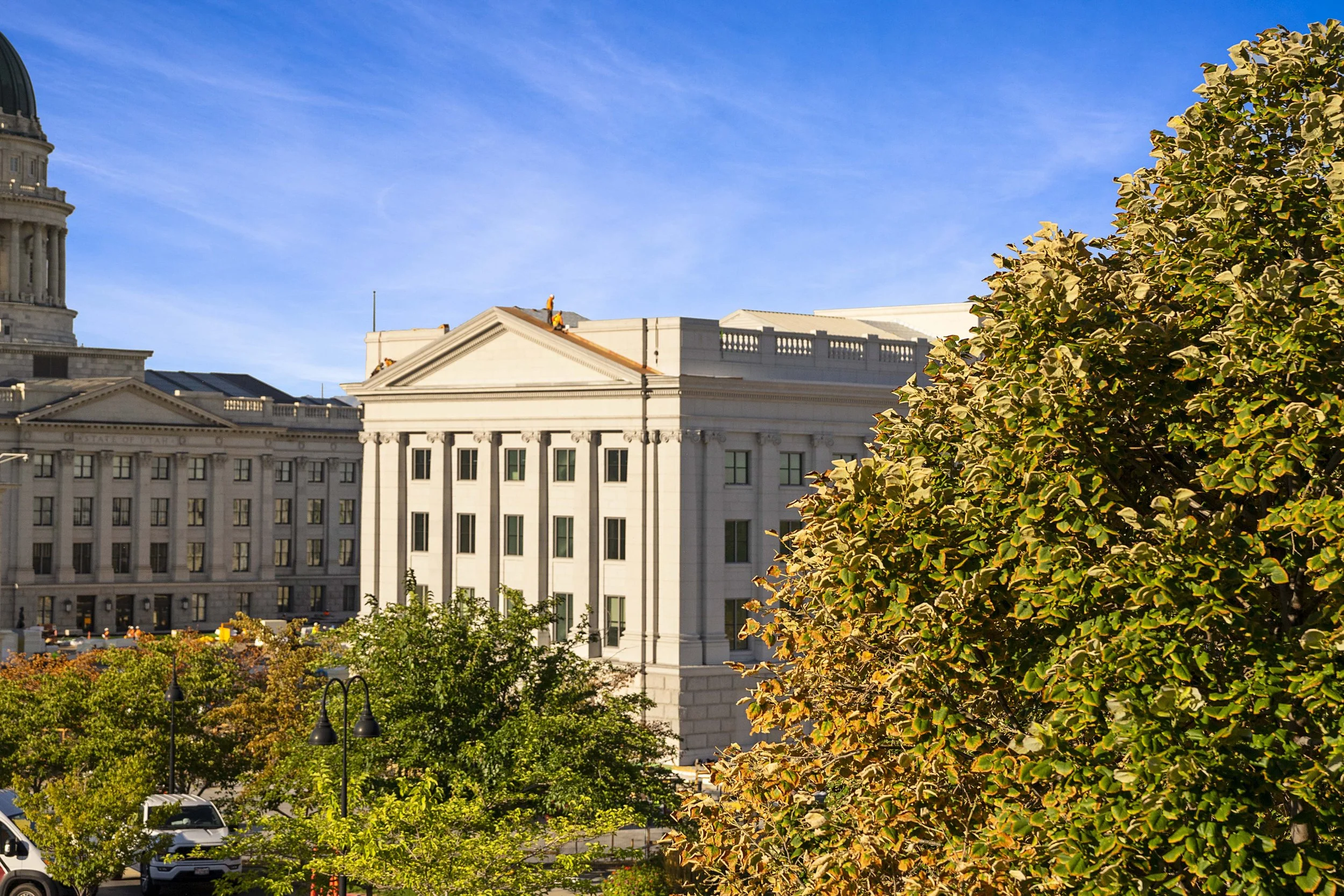 Cityscape with historic white stone government building, trees with green and yellow leaves, and clear blue sky.