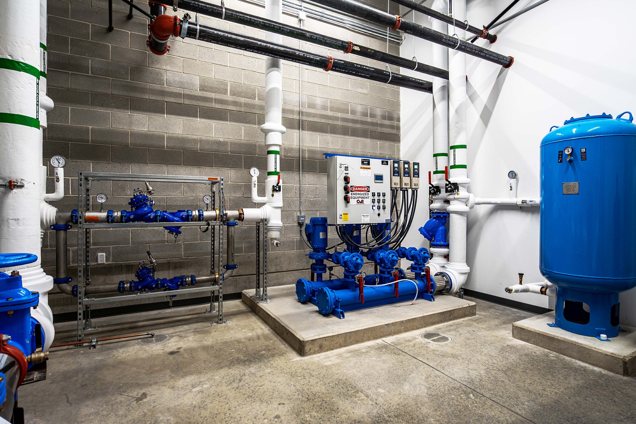 Industrial equipment and pipes, including a large blue tank and a blue pump, installed on concrete flooring in a utility room.