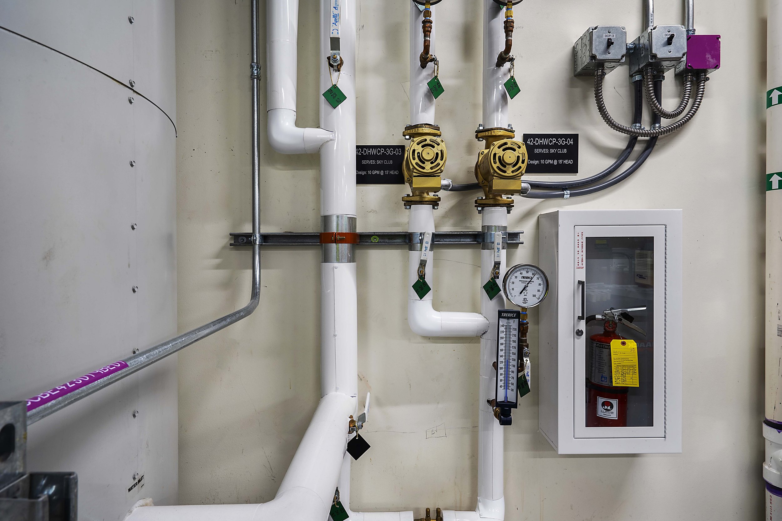 Industrial plumbing with white pipes, brass valves, pressure gauge, and electrical boxes on a beige wall. Fire extinguisher in a white cabinet to the right.