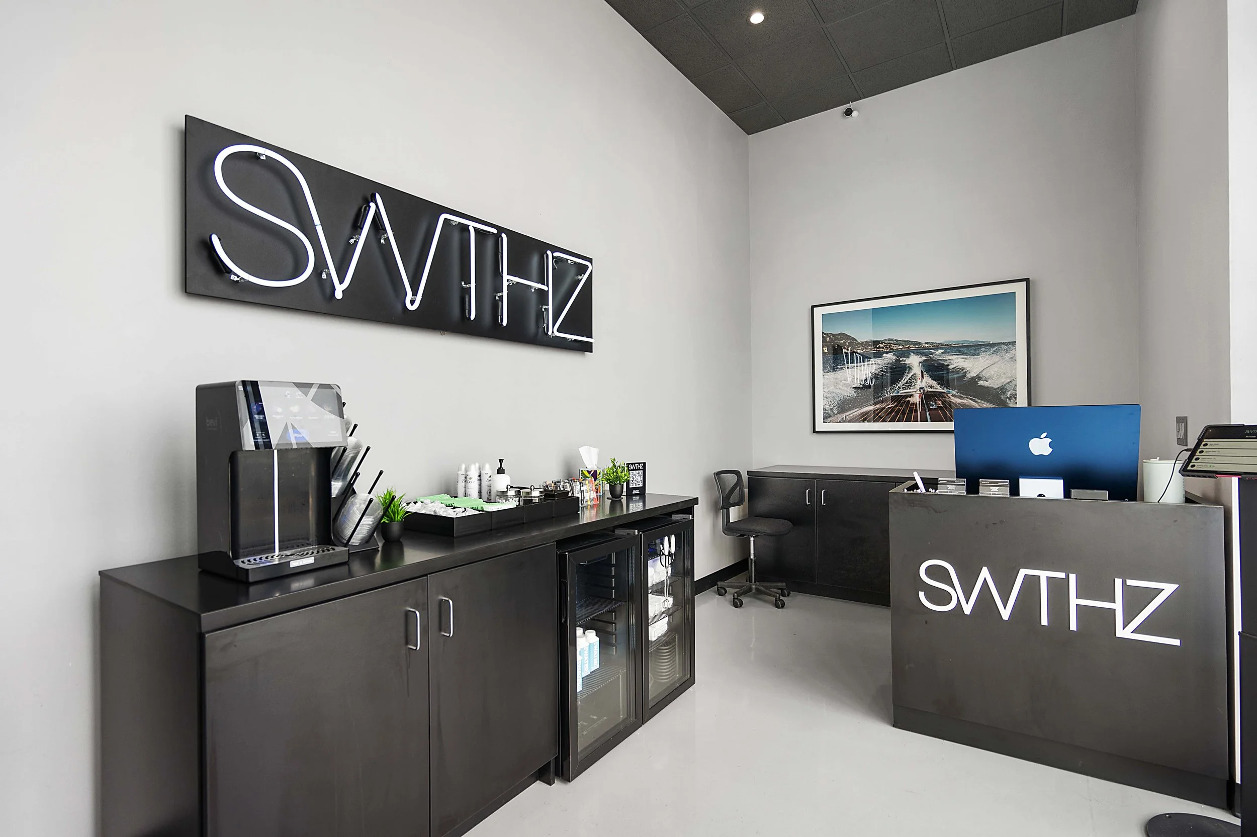 Office reception area with a dark gray counter featuring the white illuminated logo 'SWTHZ' on the front, a gray cabinet underneath, a coffee machine, and a refreshment station on top. There is a black swivel chair, a framed landscape photograph on t