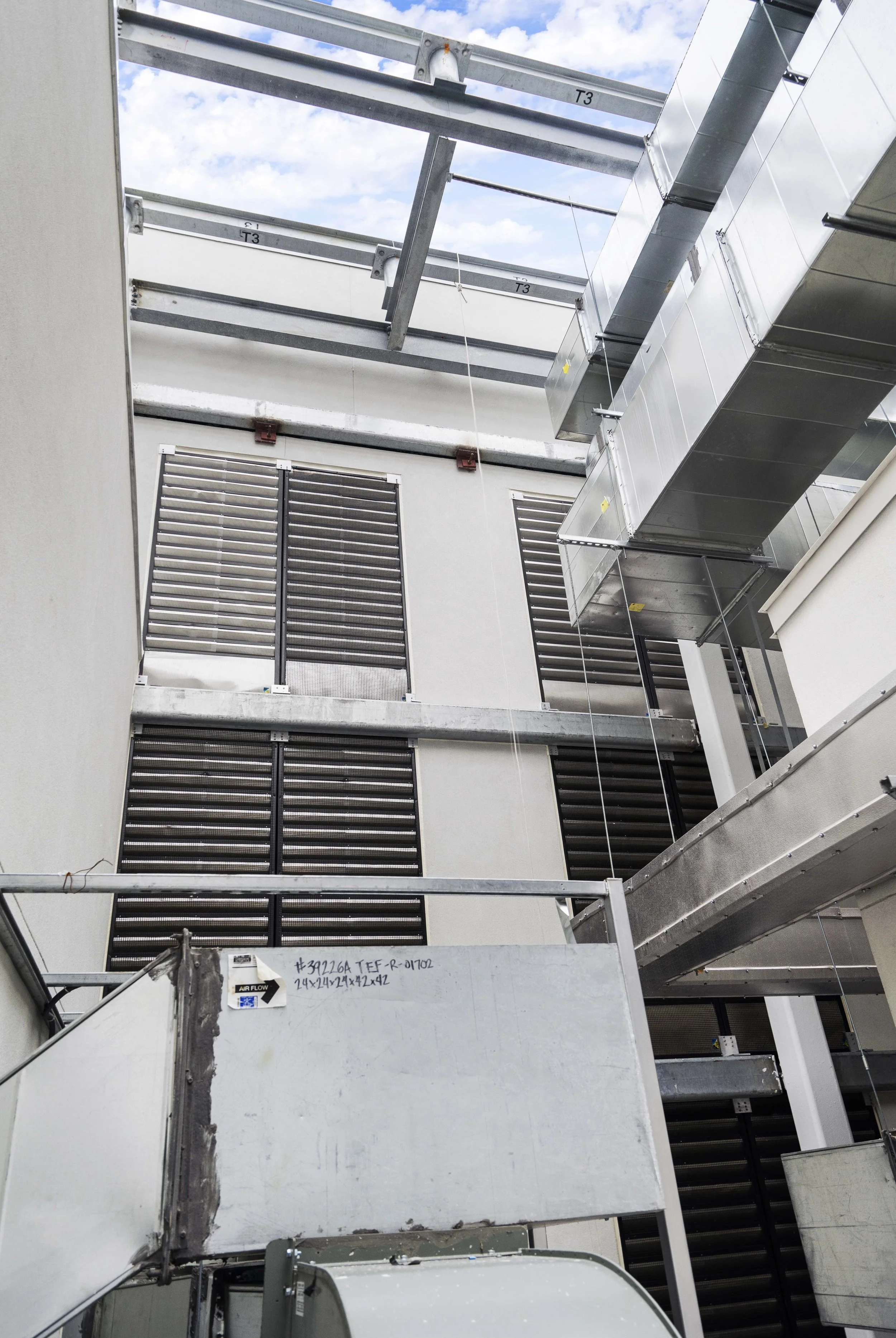An interior view of an unfinished building with exposed metal ductwork, ventilation grilles on the wall, and overhead steel beams against a partly cloudy sky.