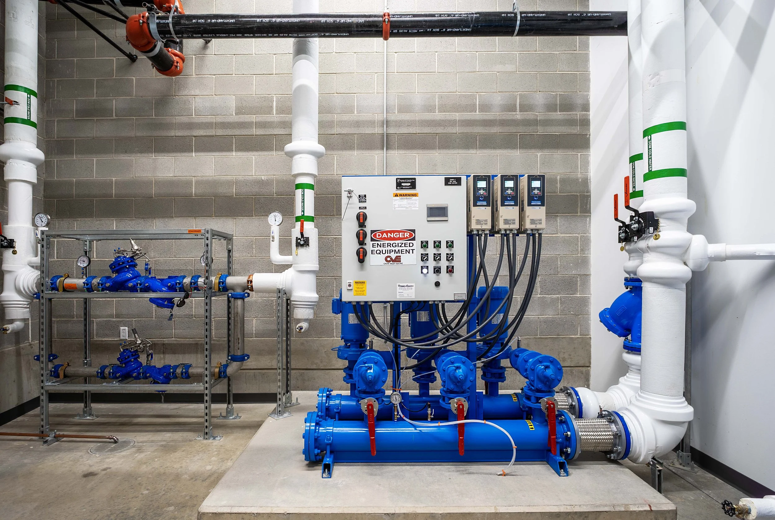 Industrial water pump system with blue pumps, white pipes, gauges, and control panel in a mechanical room.