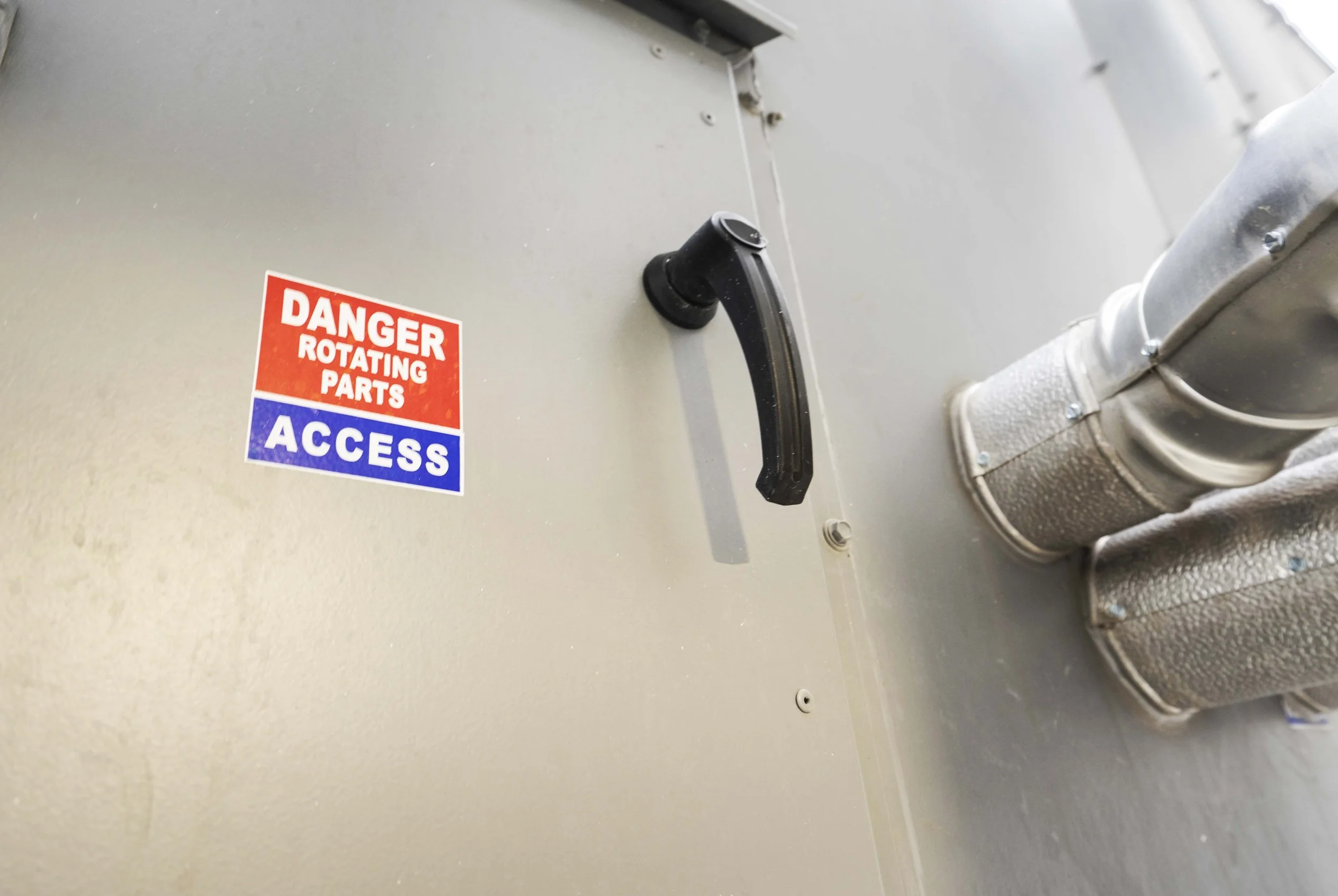 Close-up of a wall with a black handle, a ventilation pipe, and a warning sticker that reads 'Danger Rotating Parts Access' in red, white, and blue.