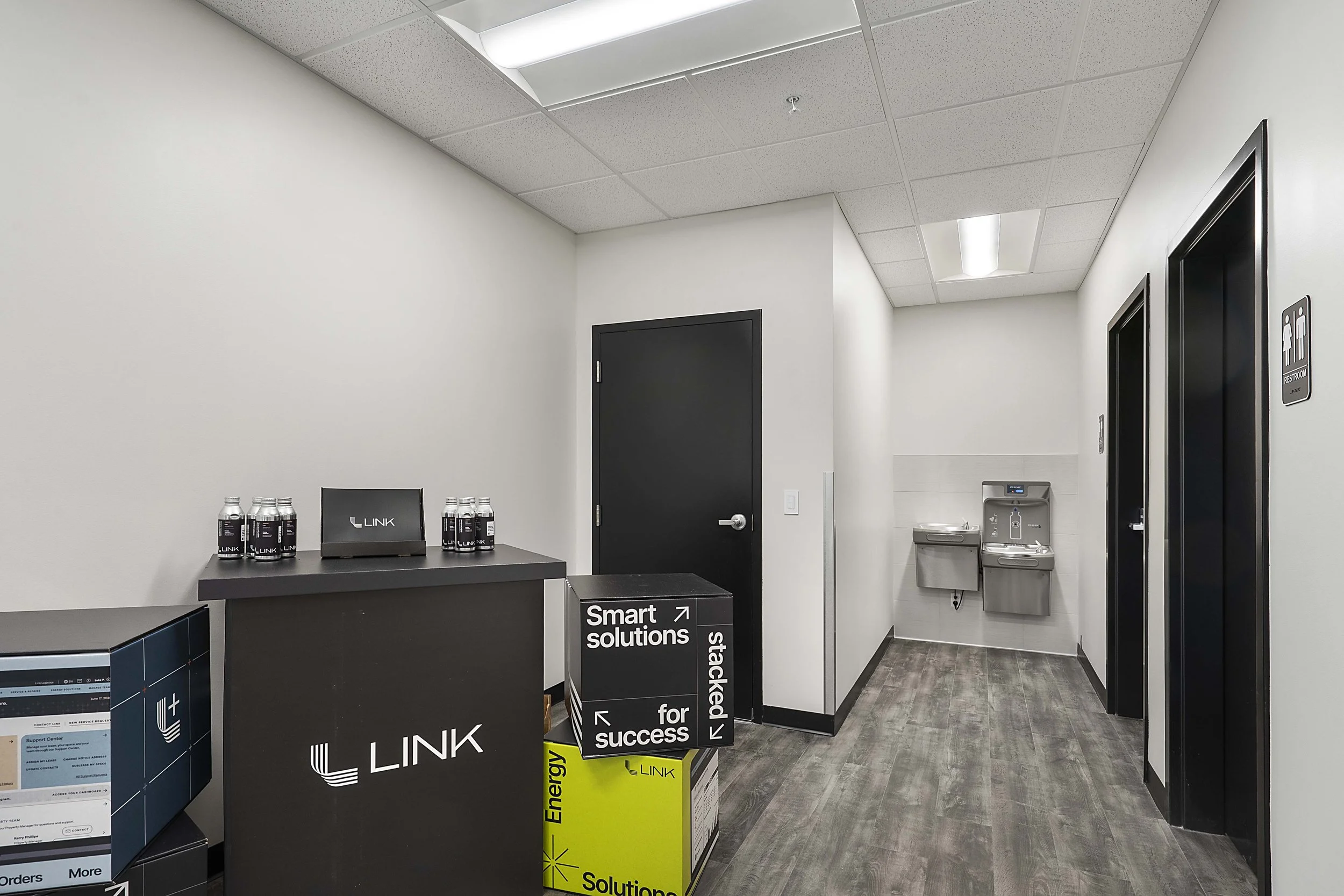 Office hallway with water fountains, black and white walls, ceiling lights, and a black reception desk with bottles and a monitor.