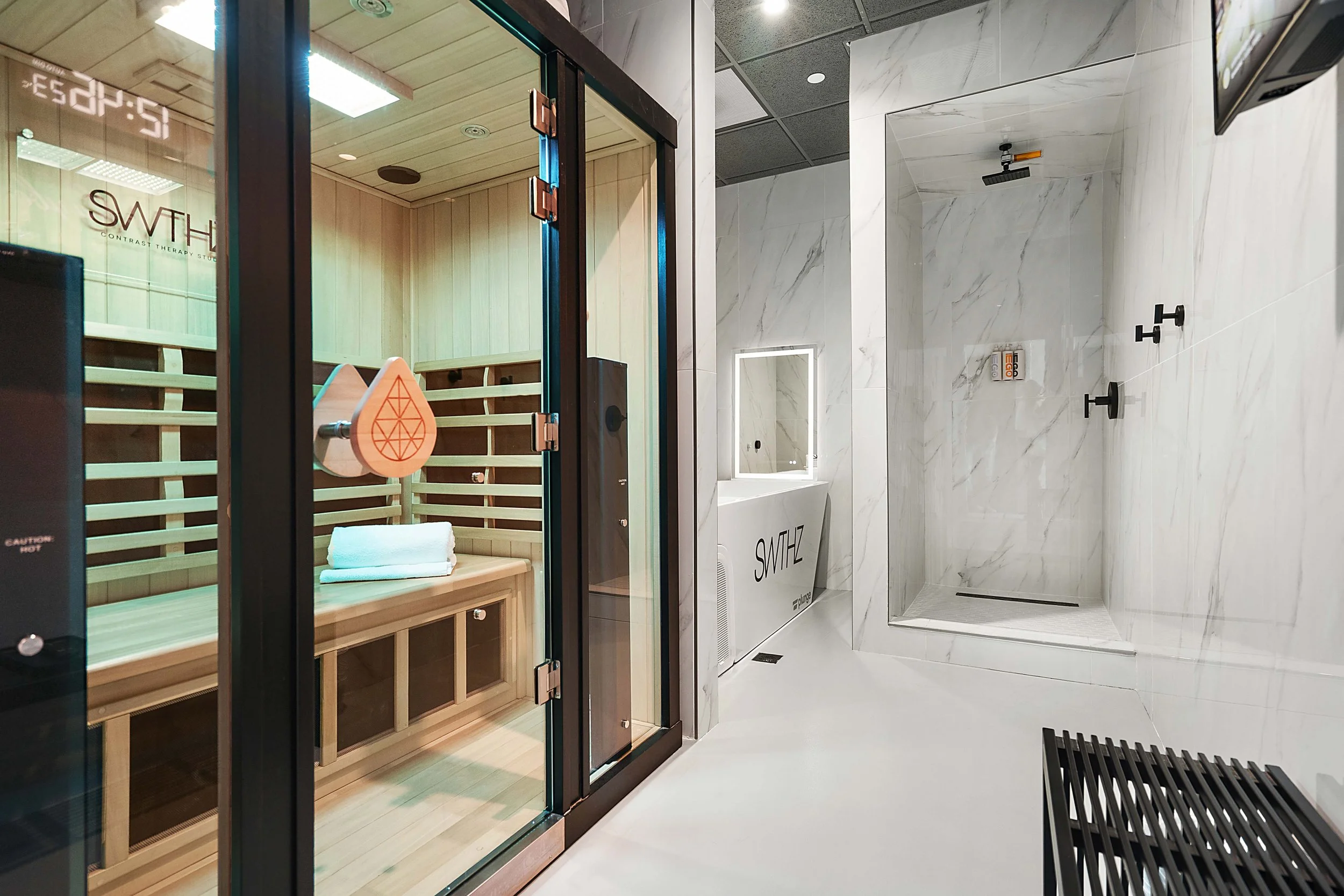 A modern wellness room with a sauna on the left, featuring wooden interior and a bench with folded towels, and a marble bathroom with a shower on the right, including three black shower hooks, a mirror, and a black vent.