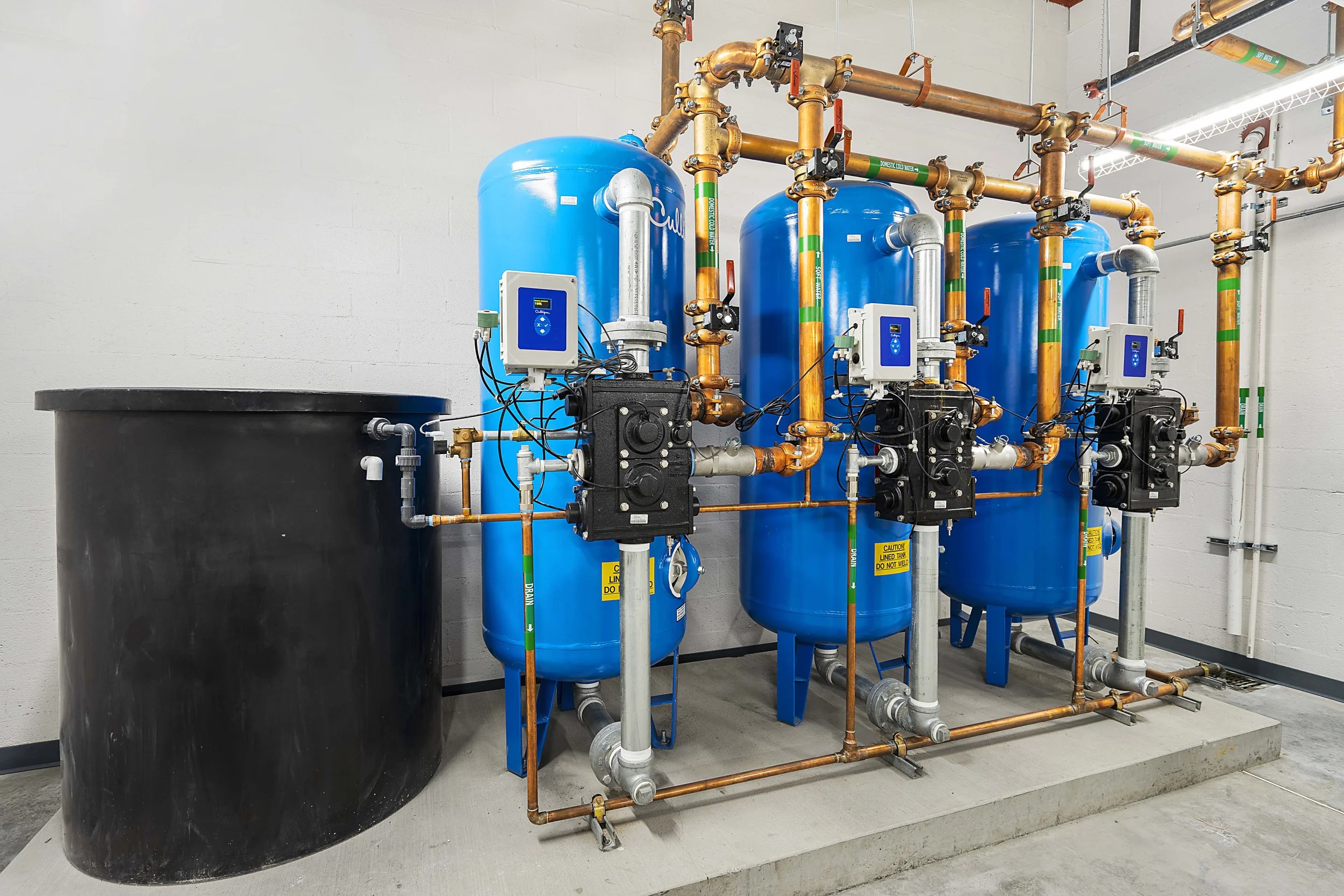Industrial water treatment system with three large blue tanks, copper pipes, and control panels in a utility room.
