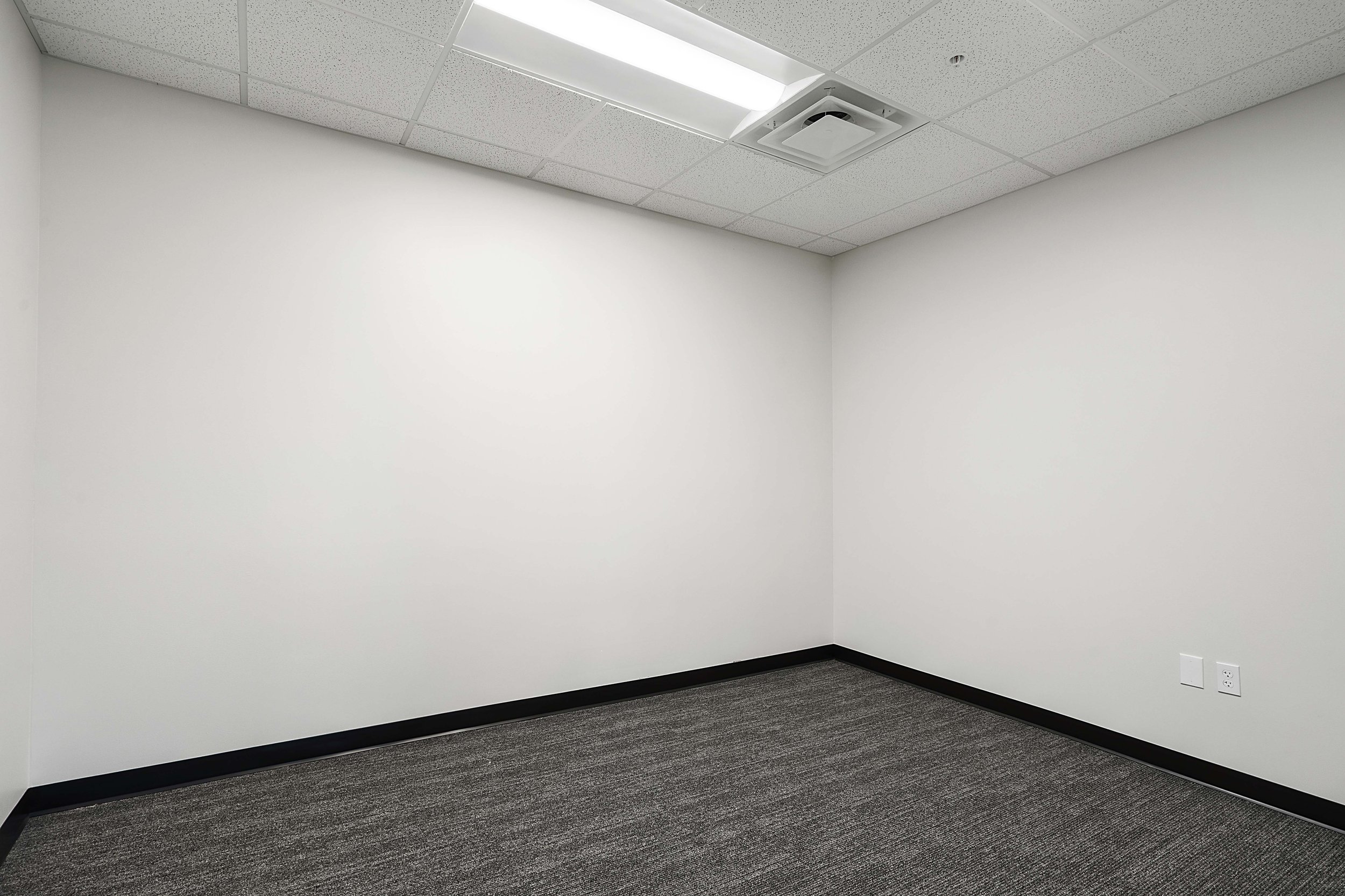 Empty conference or meeting room with white walls, gray carpet, ceiling tiles, and ceiling-mounted air conditioning vent.
