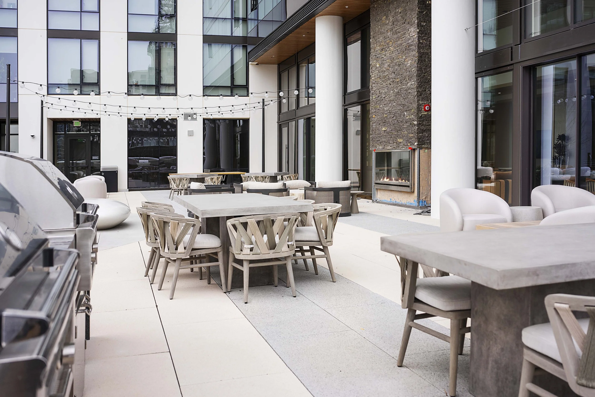 Outdoor patio area with wooden tables and chairs, white armchairs, a built-in fireplace, string lights overhead, and a modern apartment building in the background.