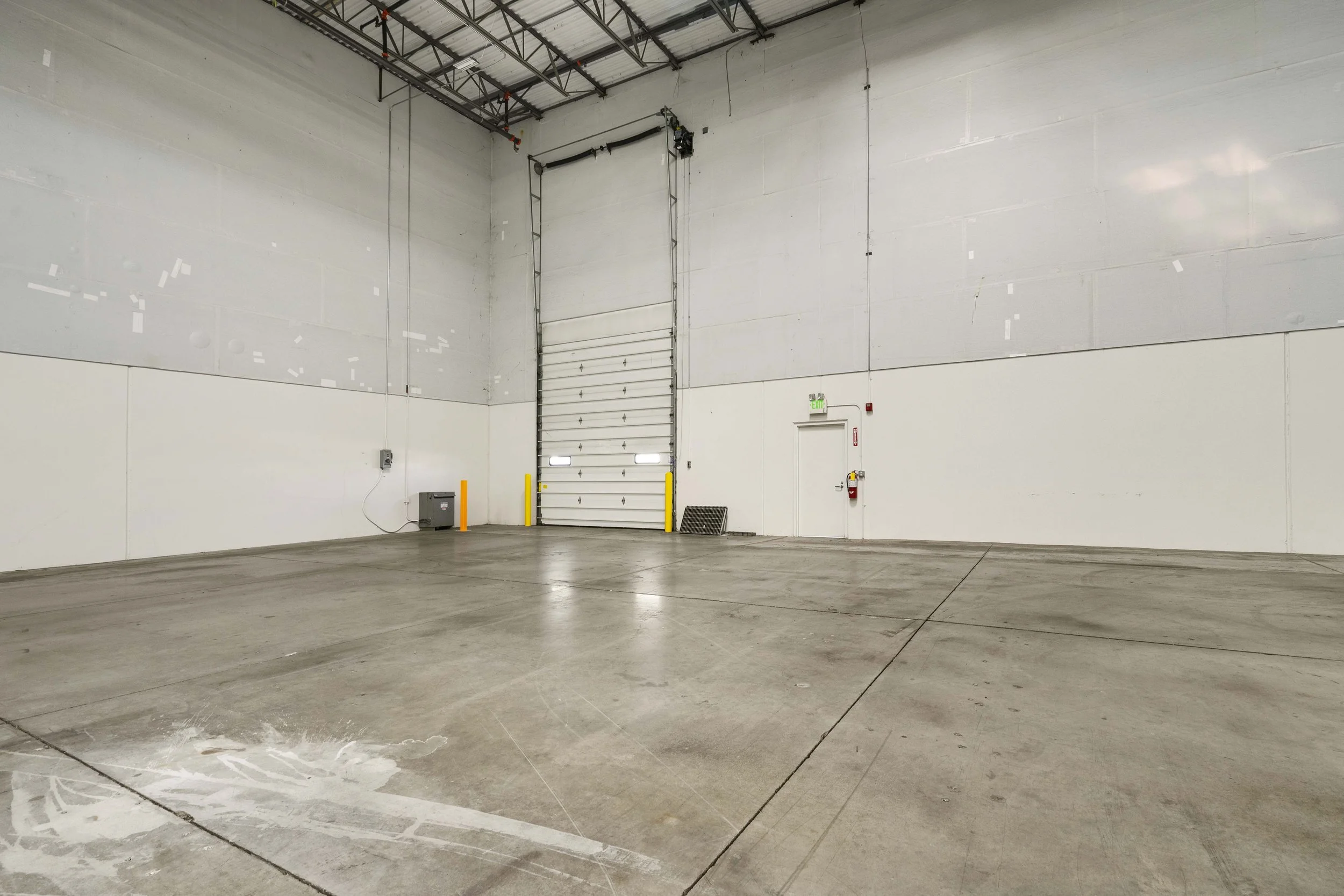 Empty warehouse with concrete floor, white walls, industrial door, and a small door with an emergency exit sign.