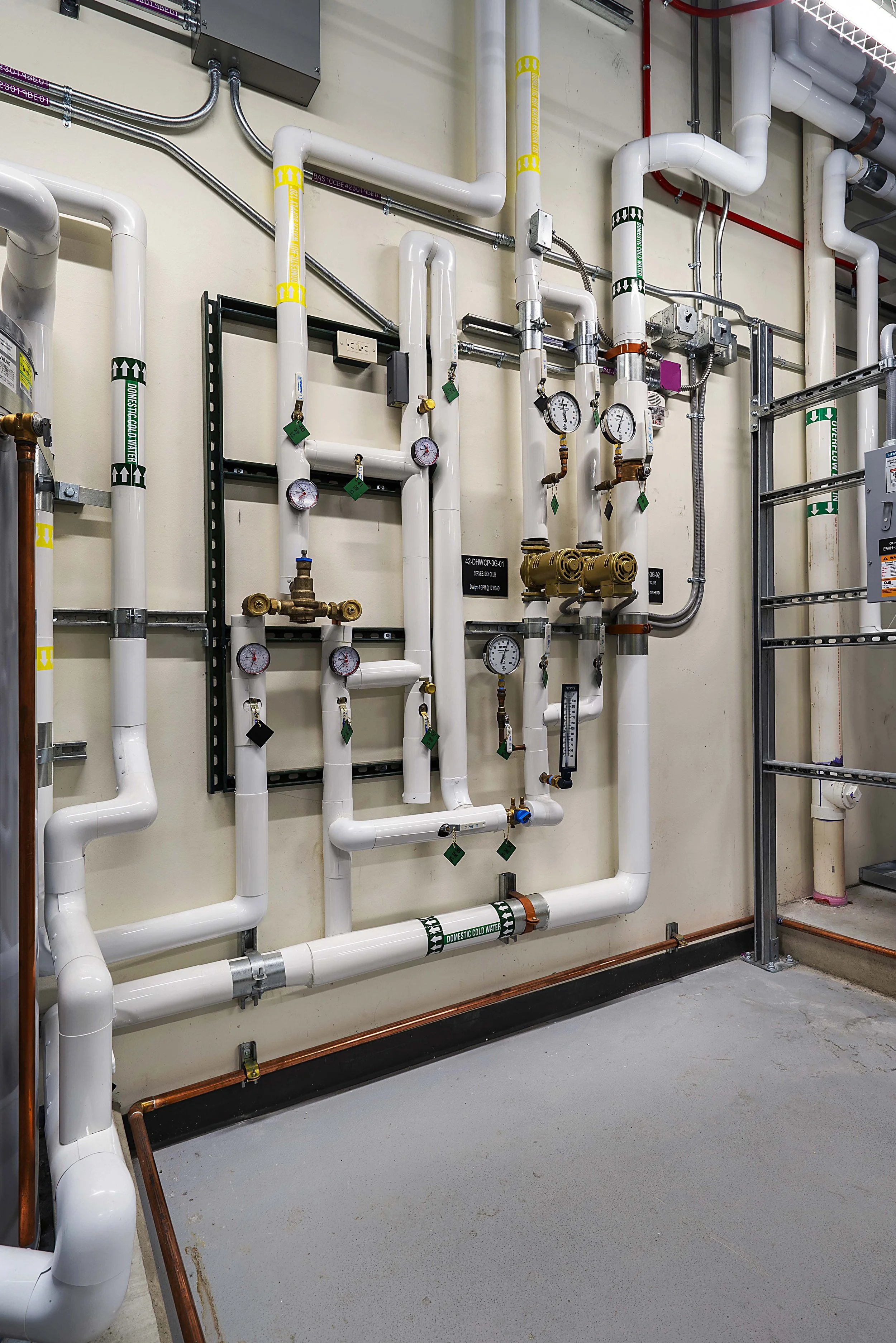 An engineered plumbing system with white pipes, pressure gauges, and valves on a wall, with some copper pipes and electrical conduit visible.