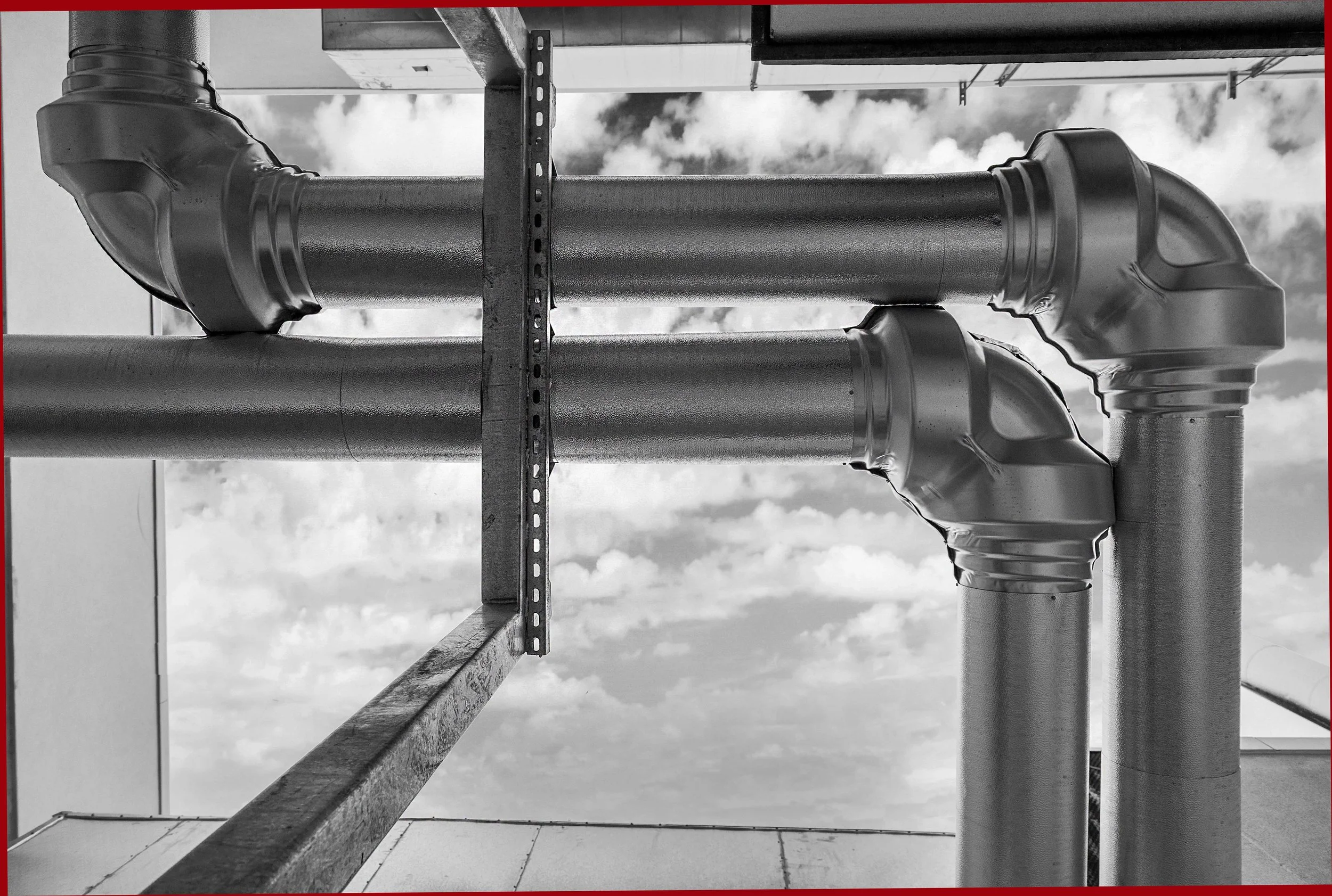 Black and white photo of industrial metal pipes with elbows, mounted on a rooftop under a cloudy sky.