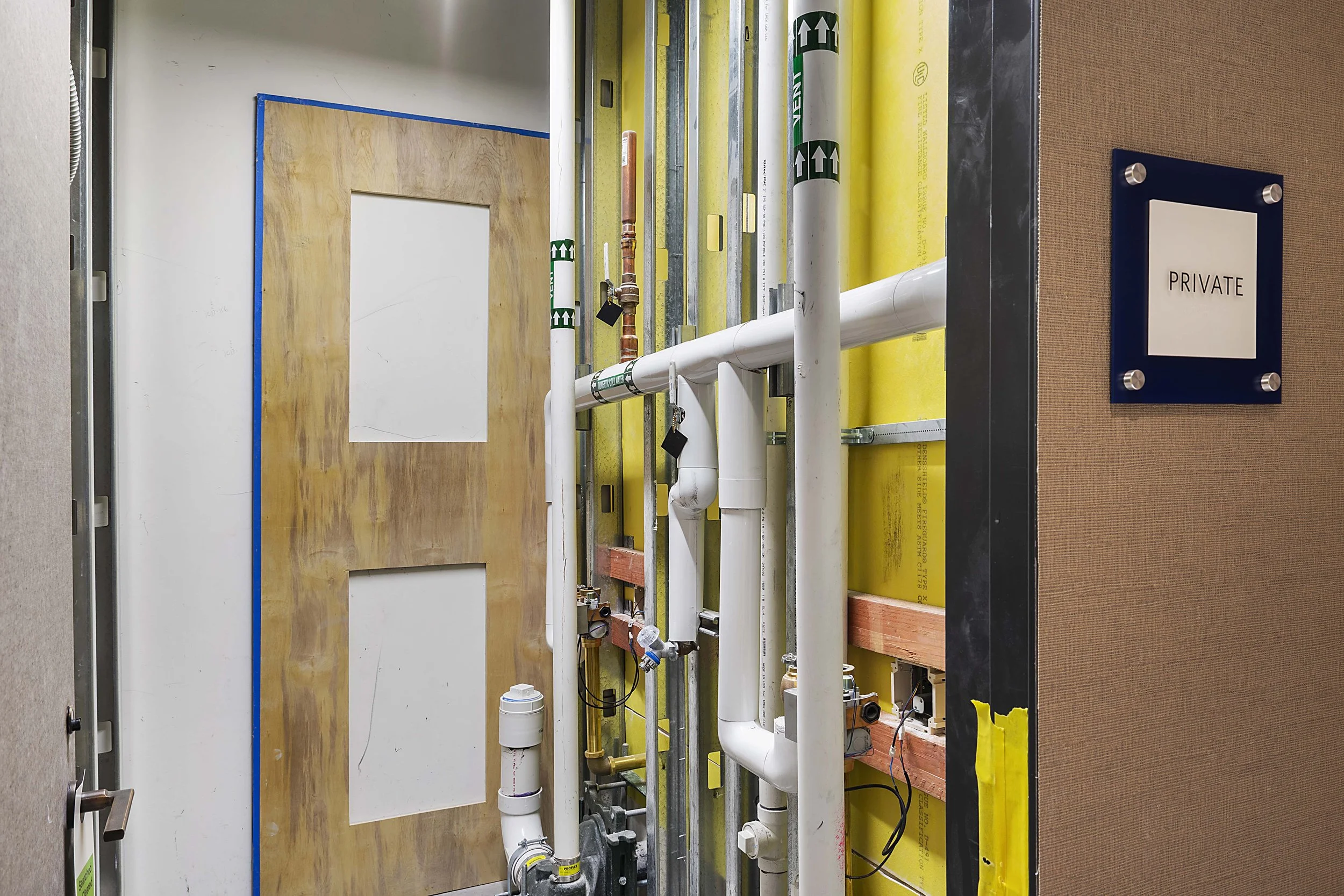 Construction or plumbing area behind a temporary door with pipe work and a 'Private' sign on the wall.