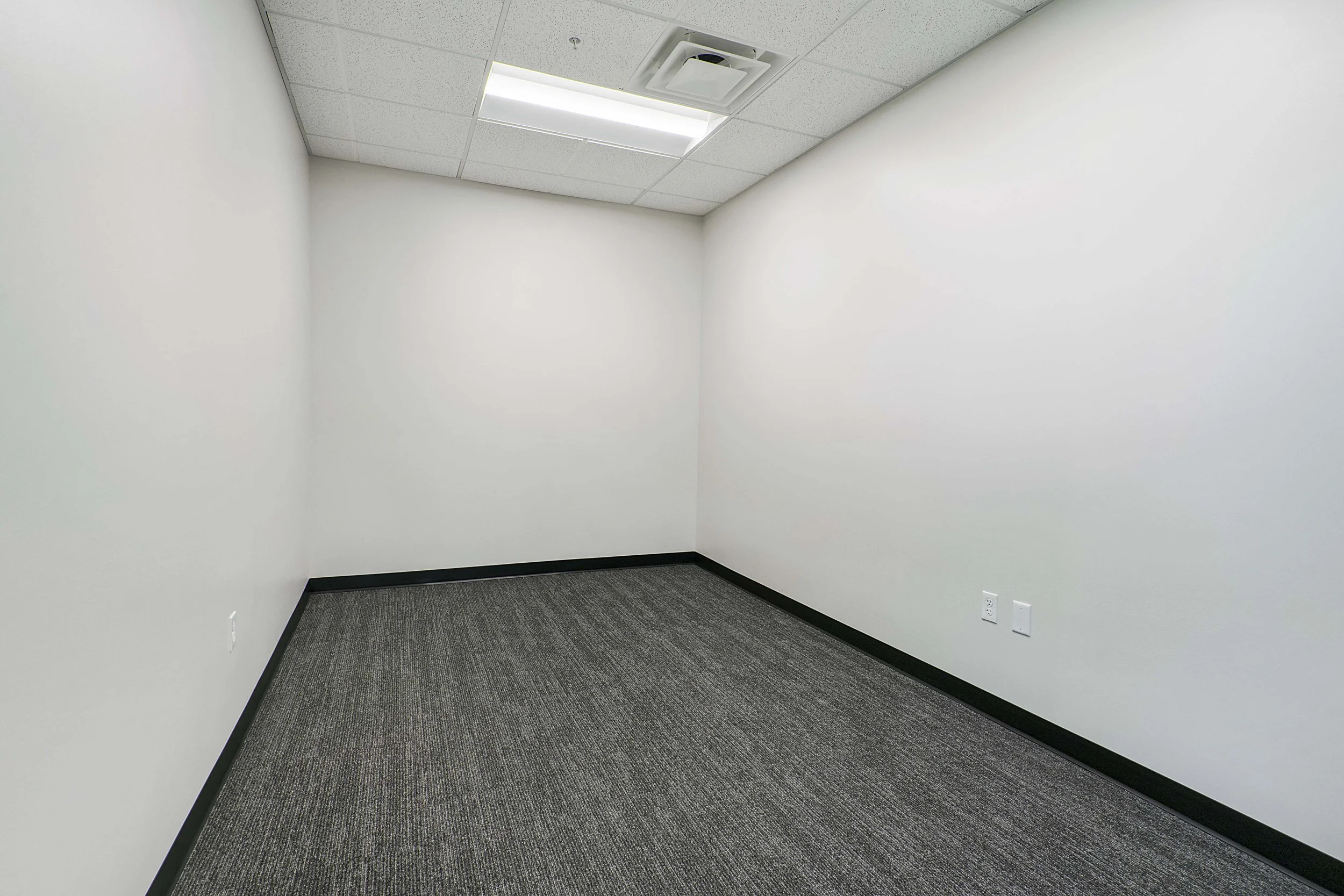 Empty office room with white walls, gray carpet, ceiling with fluorescent light, and ceiling vent.