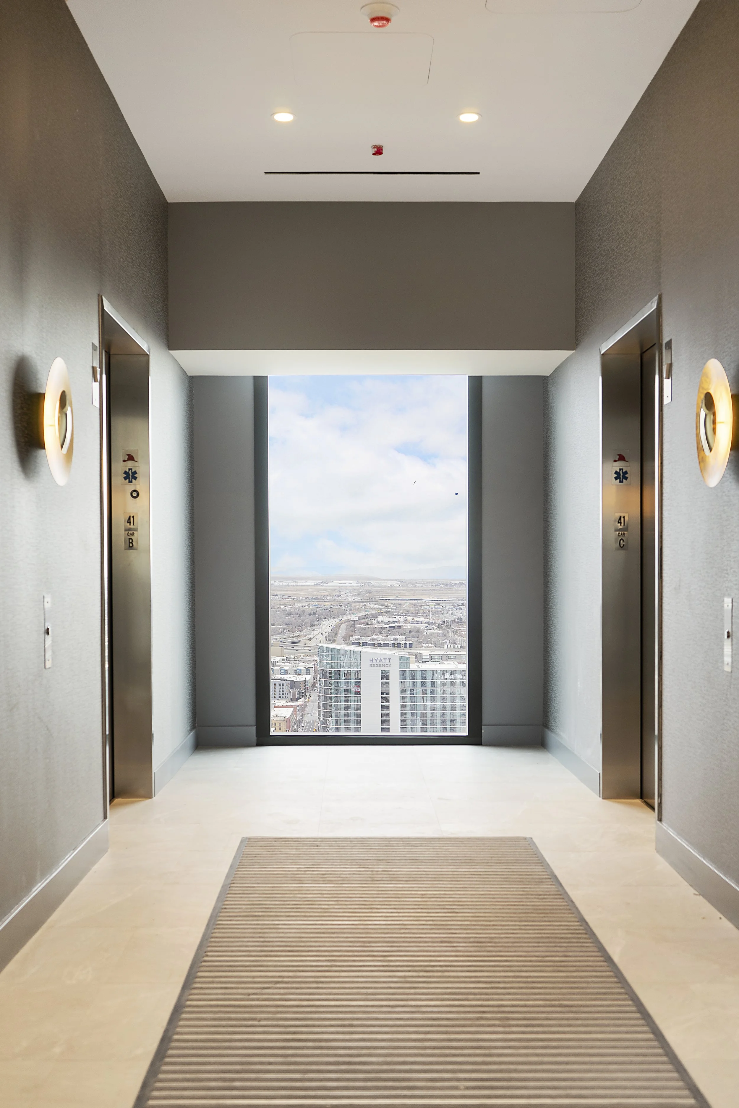 Two elevators in a modern hotel corridor with a large window overlooking a cityscape and cloudy sky.