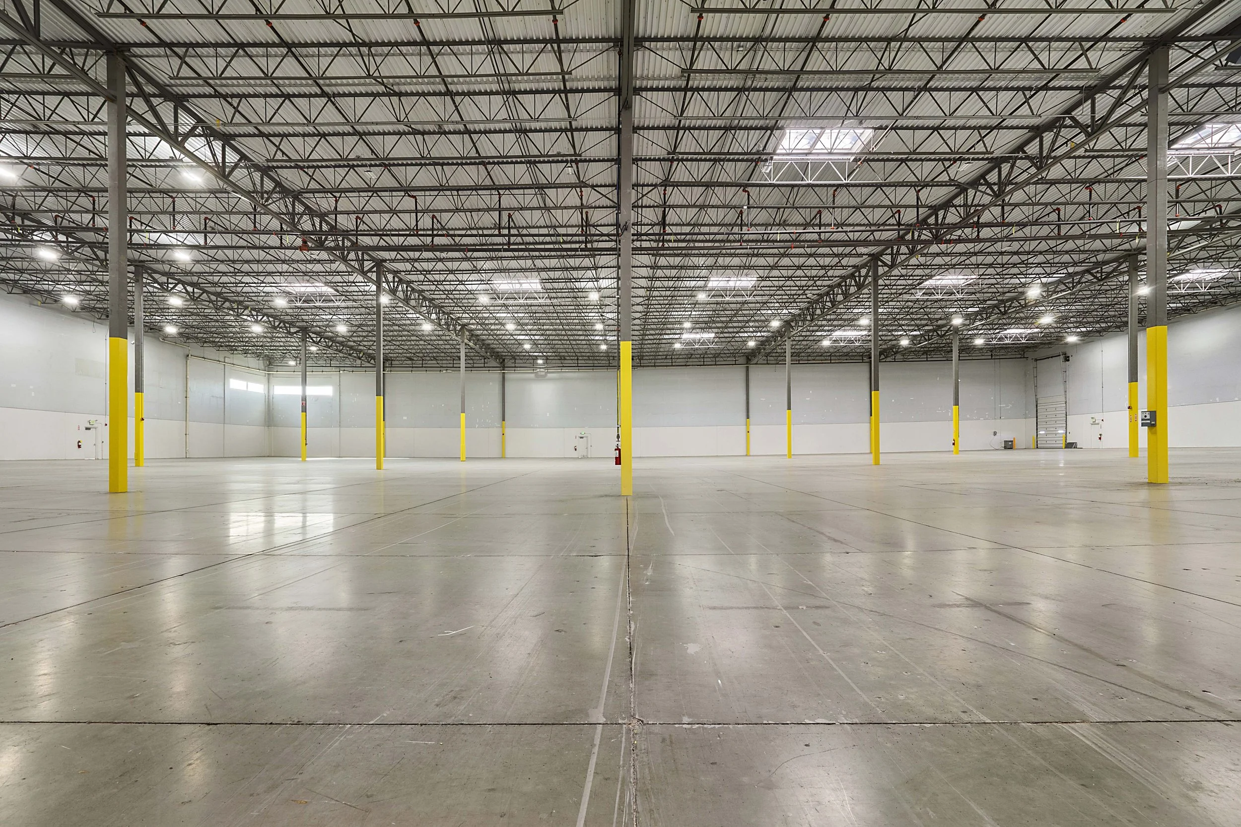 Empty warehouse with high ceiling, metal framework, and bright overhead lights.