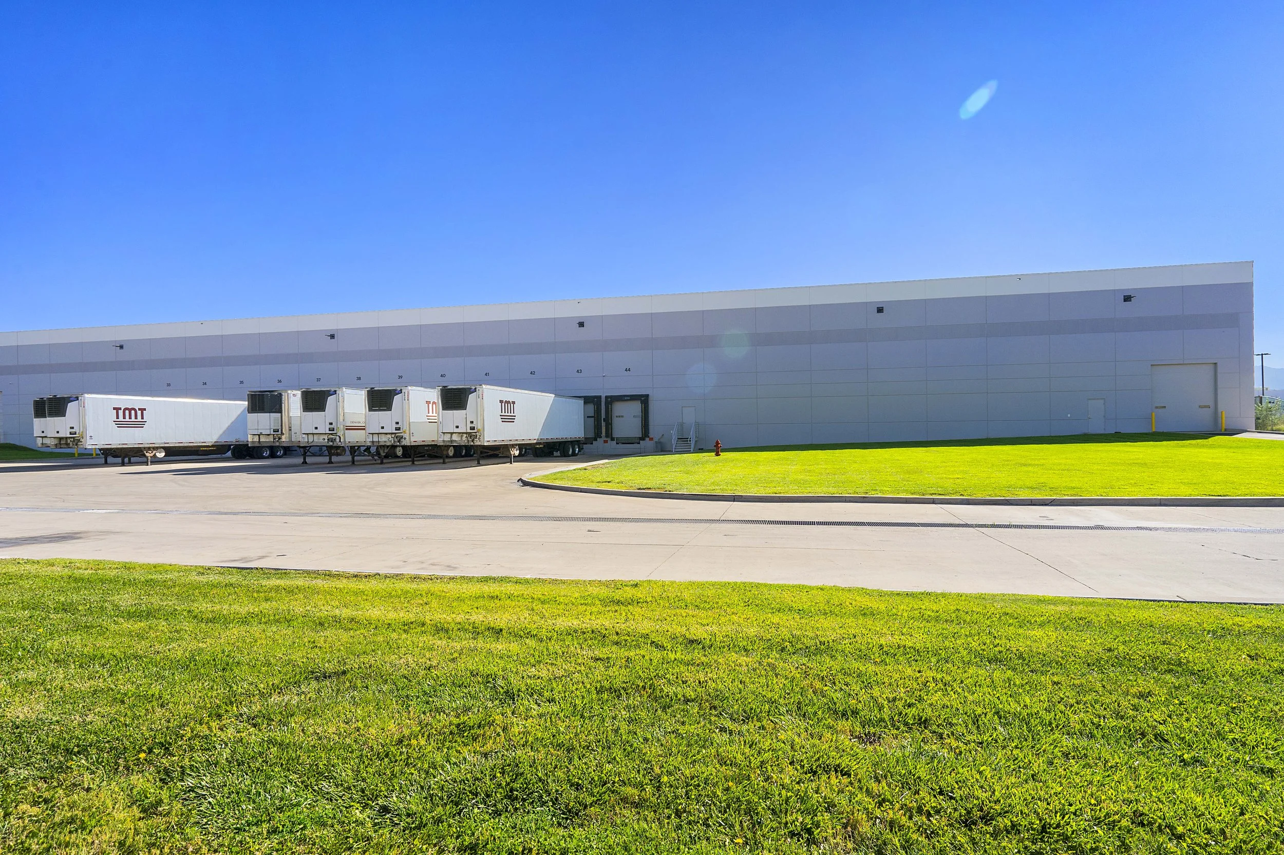 White warehouse with several parked trucks, a large door, and a green grassy area in the foreground.