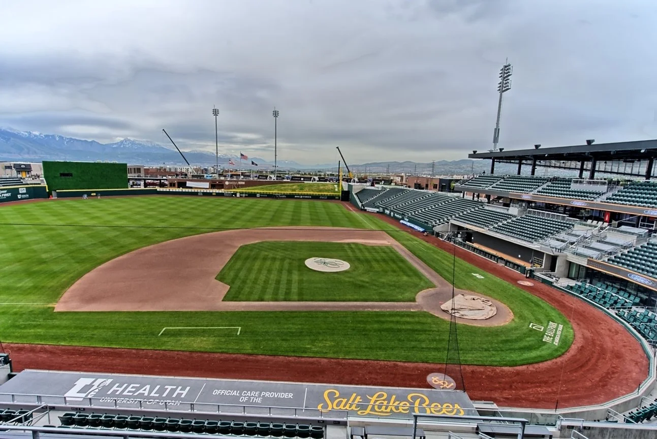 Empty baseball stadium with a field, seating areas, and city view with mountains in the background.
