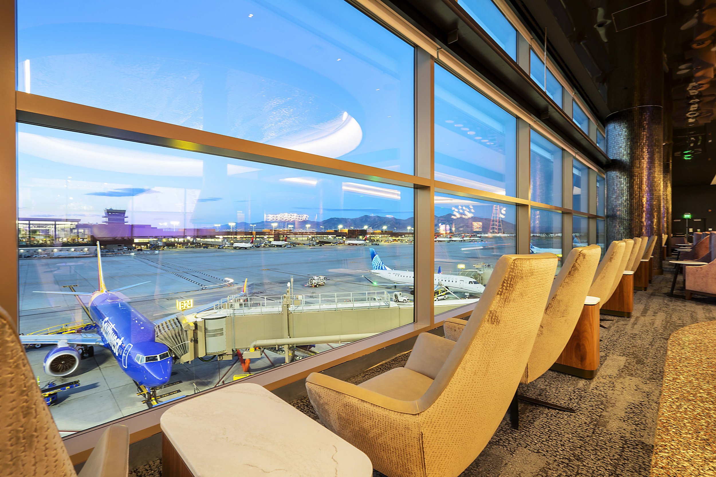 Airport terminal lounge with large windows overlooking parked airplanes on the tarmac, some with jet bridges attached, and mountains in the distance; row of beige lounge chairs along the window with a patterned carpet on the floor.