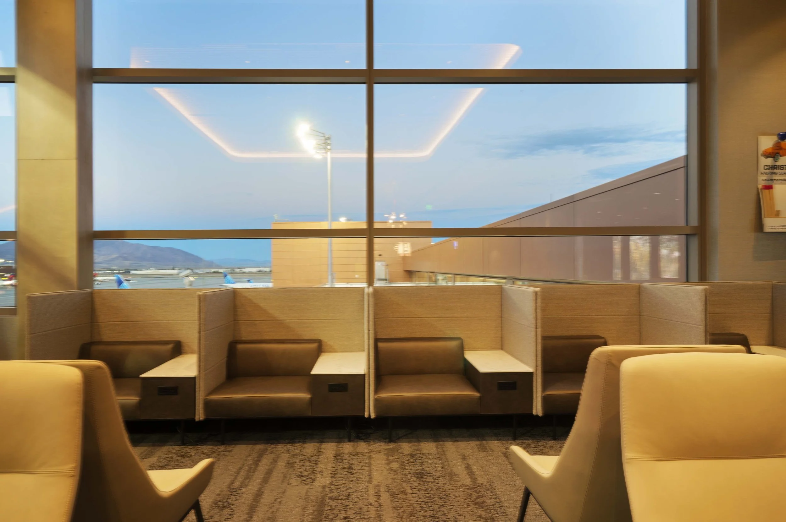 View of an airport gate area with beige seating and a large window showing planes on the tarmac and mountains in the distance during dusk or dawn.