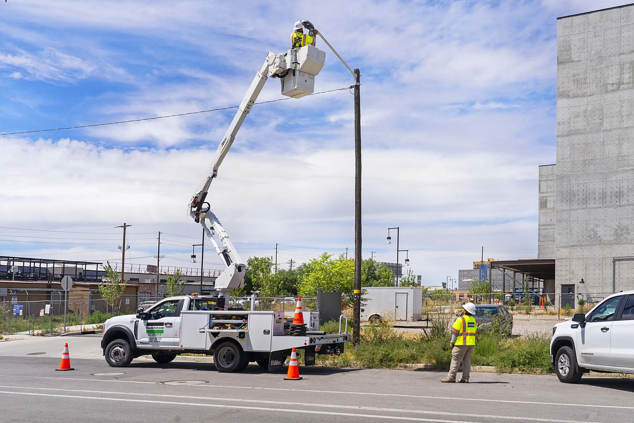 A worker in a safety vest and helmet is elevated in a bucket lift attached to a white utility truck, fixing or inspecting a streetlight or utility pole.
