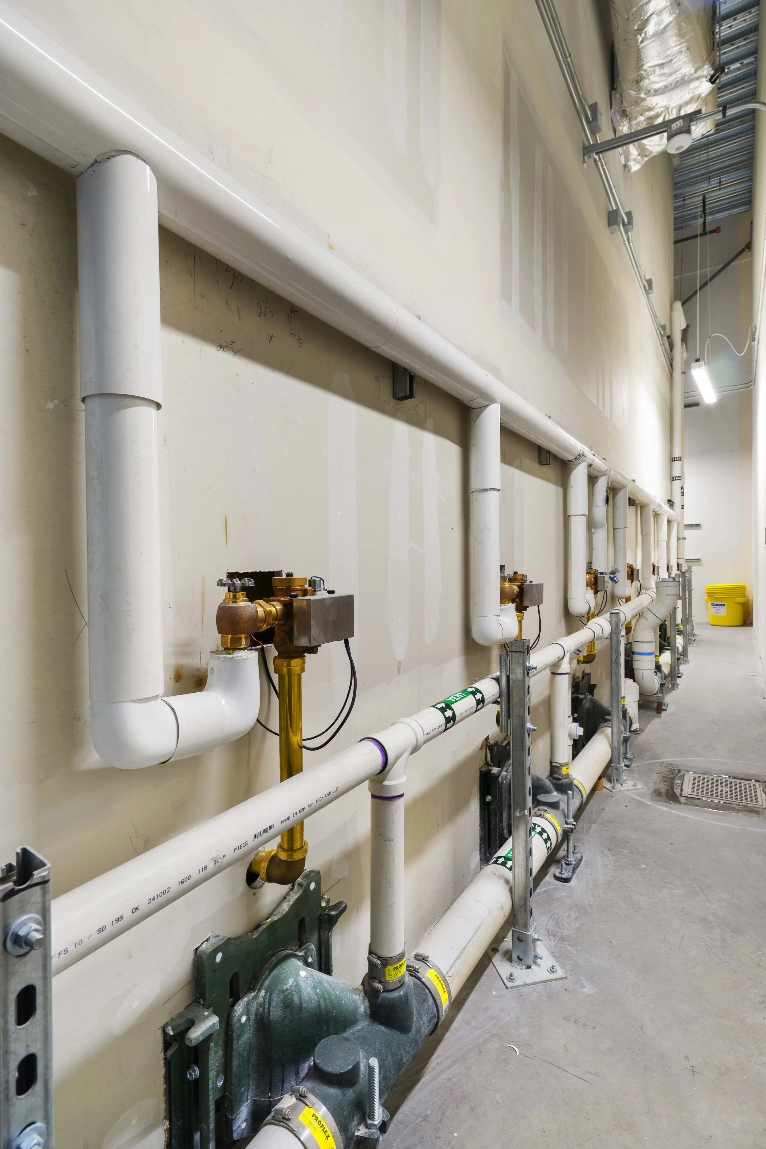 Internal view of a room with installed plumbing pipes, valves, and brackets, with a yellow bucket in the background.