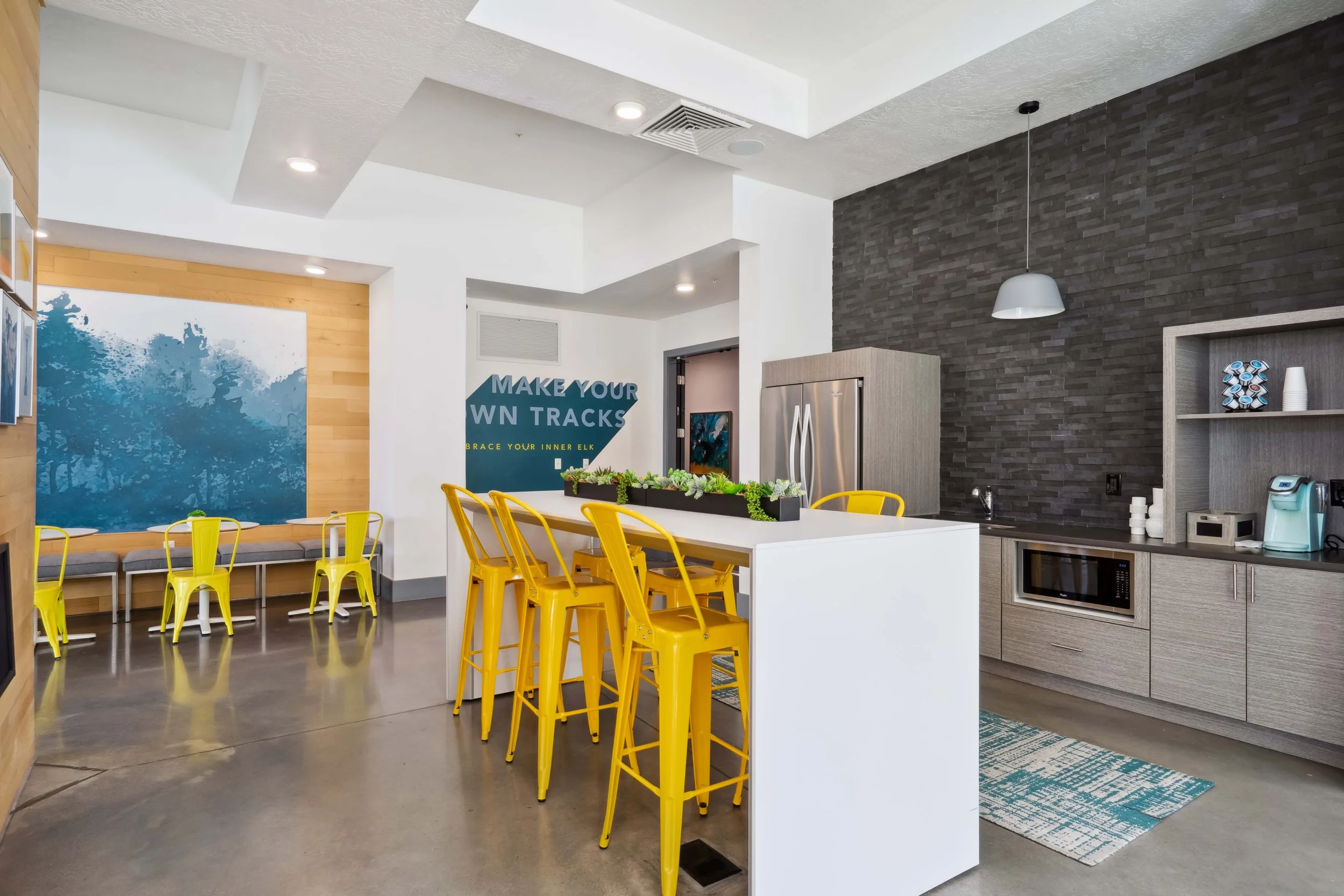 Modern kitchen and dining area with yellow barstools at a white island, gray cabinetry, a black brick accent wall, and a wall painting with trees, with a sign that says 'Make your own tracks' and 'Embrace your inner elk'.