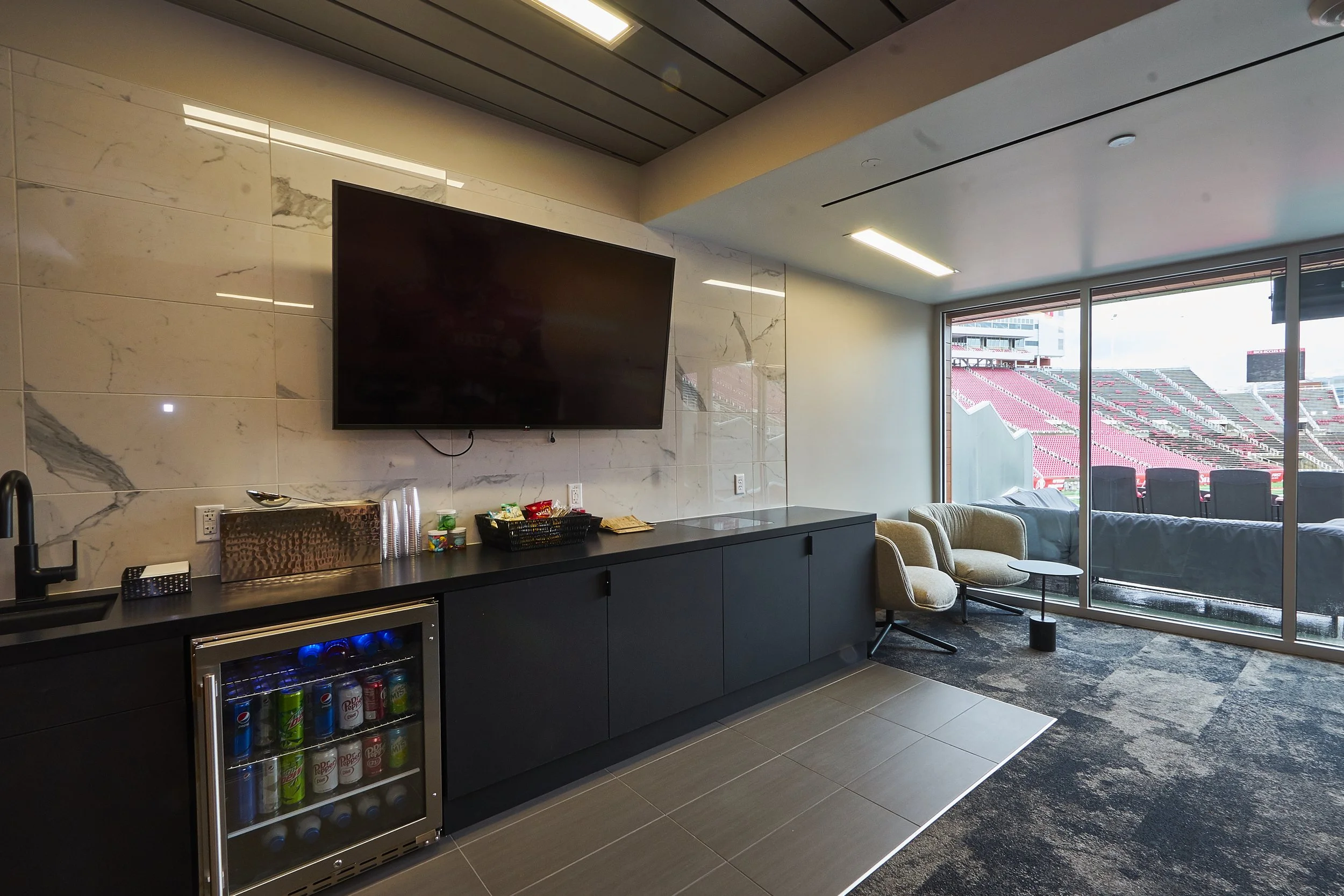 Luxury lounge area with a large TV mounted on a marble wall, a black cabinet with snacks and drinks, beige chairs, and a view of a stadium through glass doors.