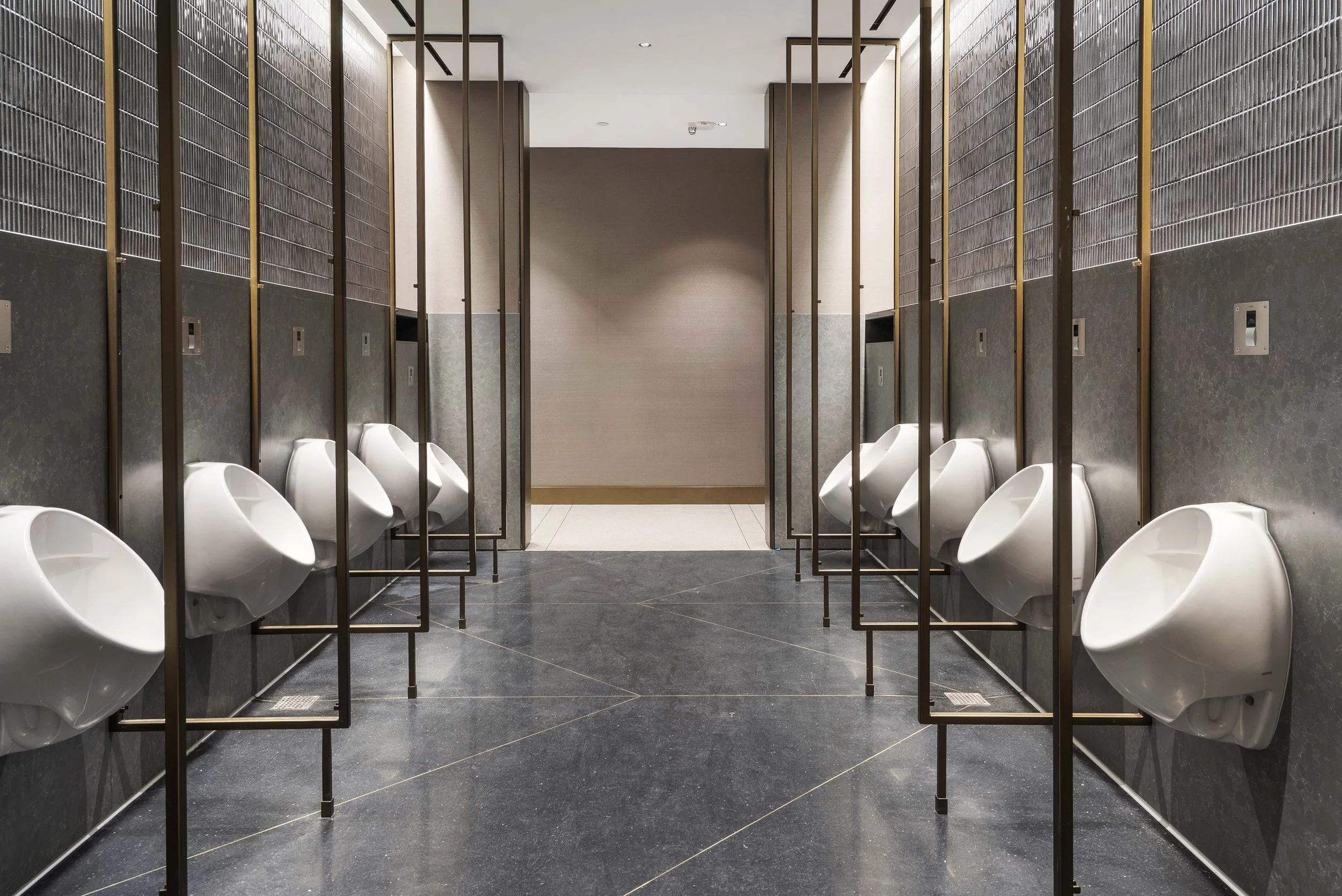 Men's public urinals in a modern, clean restroom with dark tiled floor and walls, separated by metal dividers.