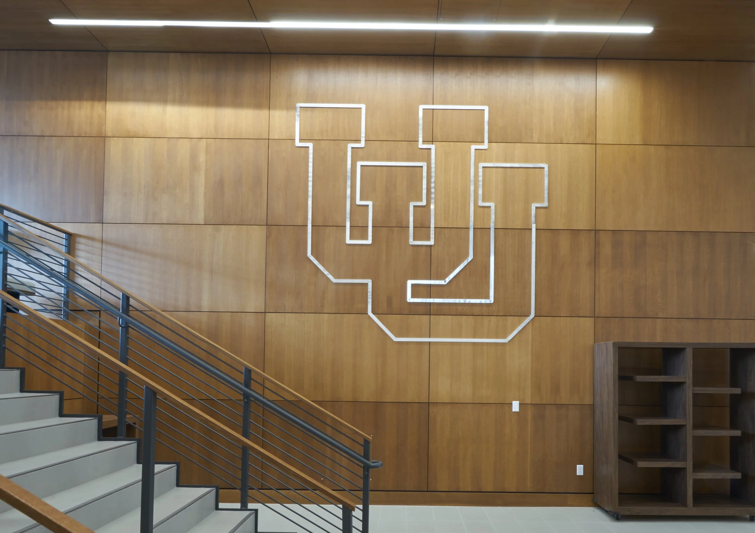 Wood-paneled wall with a large white outlined letter U installed on it. Metal staircase with wooden handrail on the left side, and empty wooden shelving on the right side.