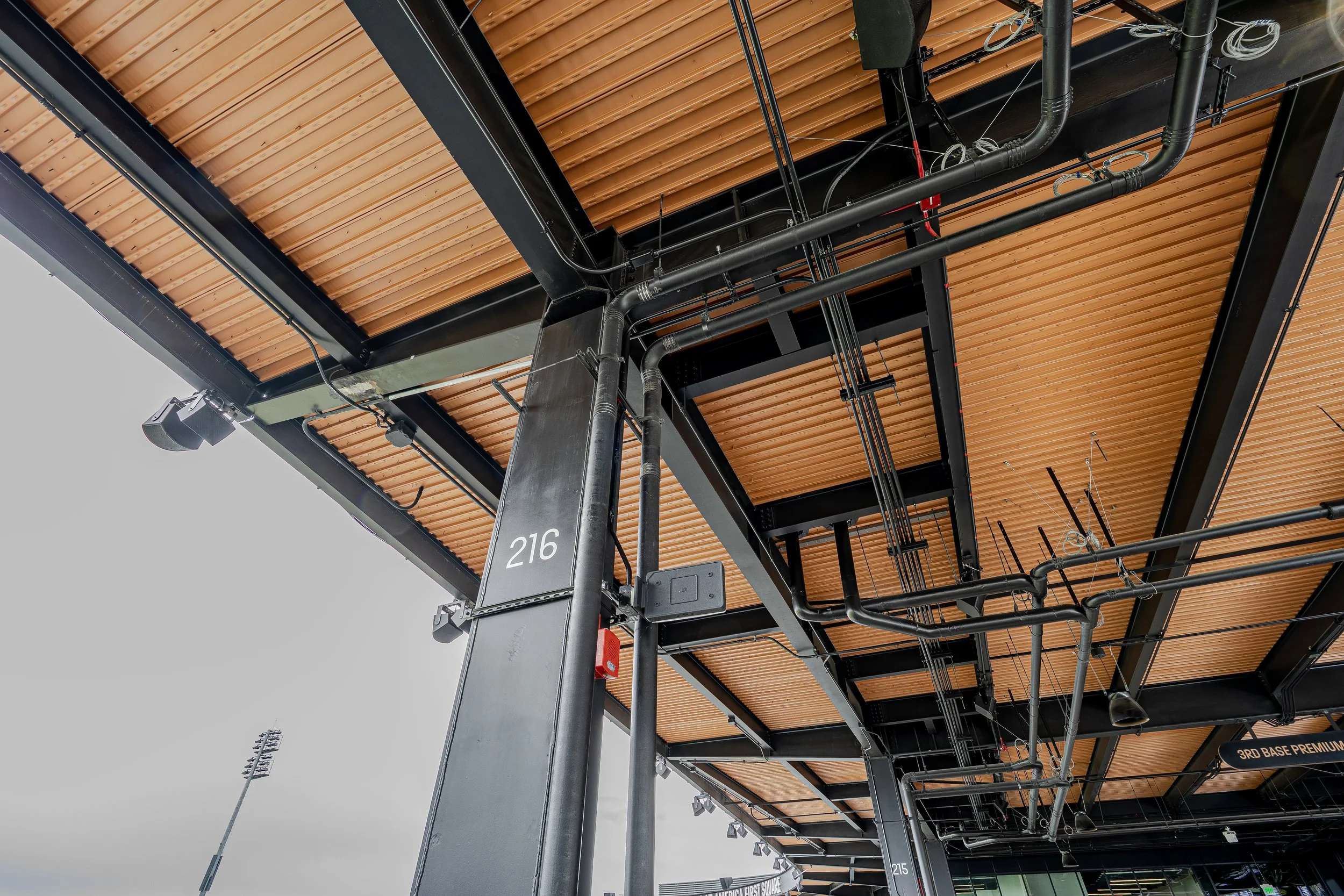 Close-up of the underside of a stadium seating area showing black steel supports, orange wooden paneling on the ceiling, and electrical conduit piping.