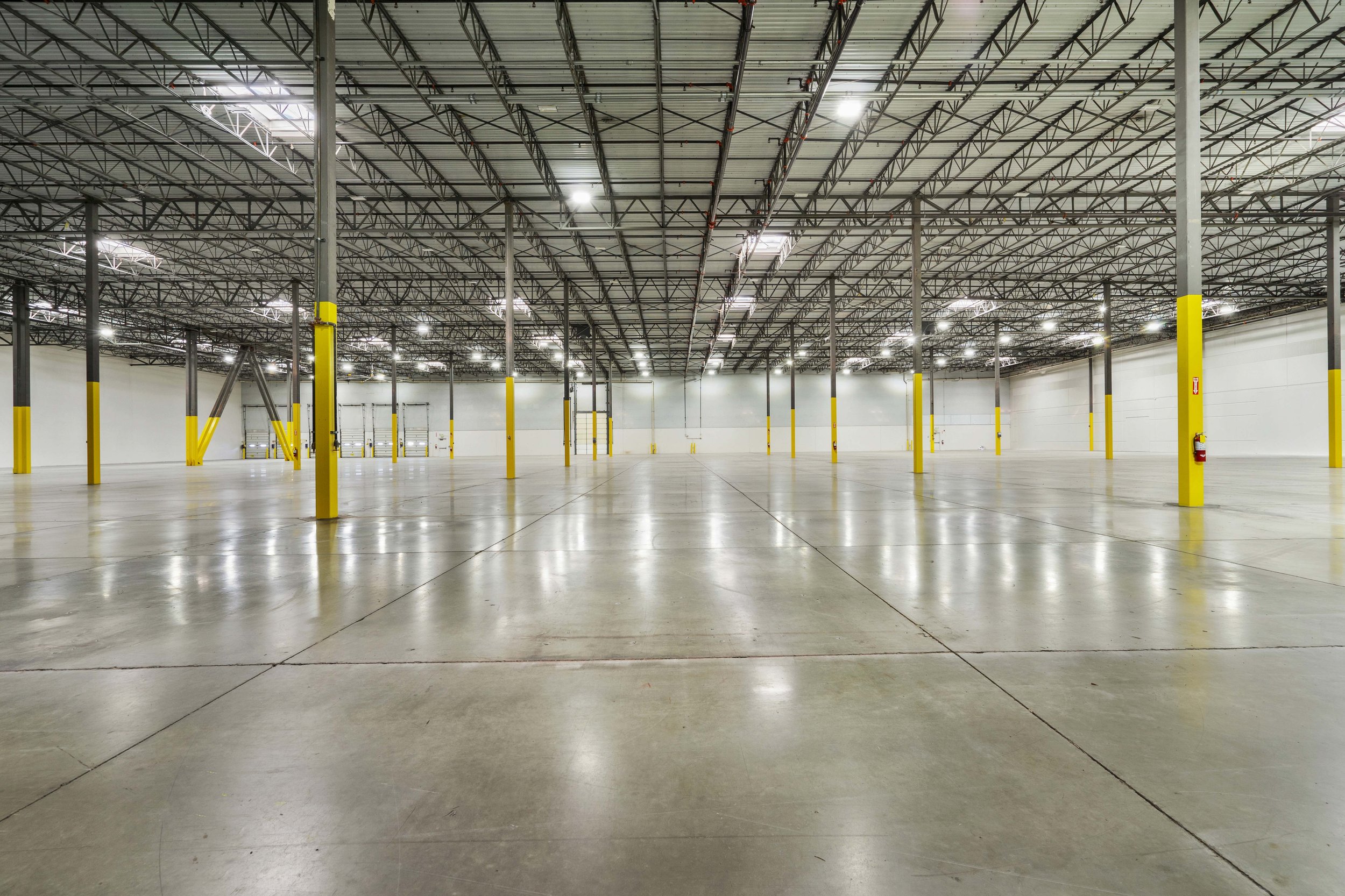 Empty warehouse with polished concrete floors, yellow safety posts, metal roof structure, and overhead lighting.