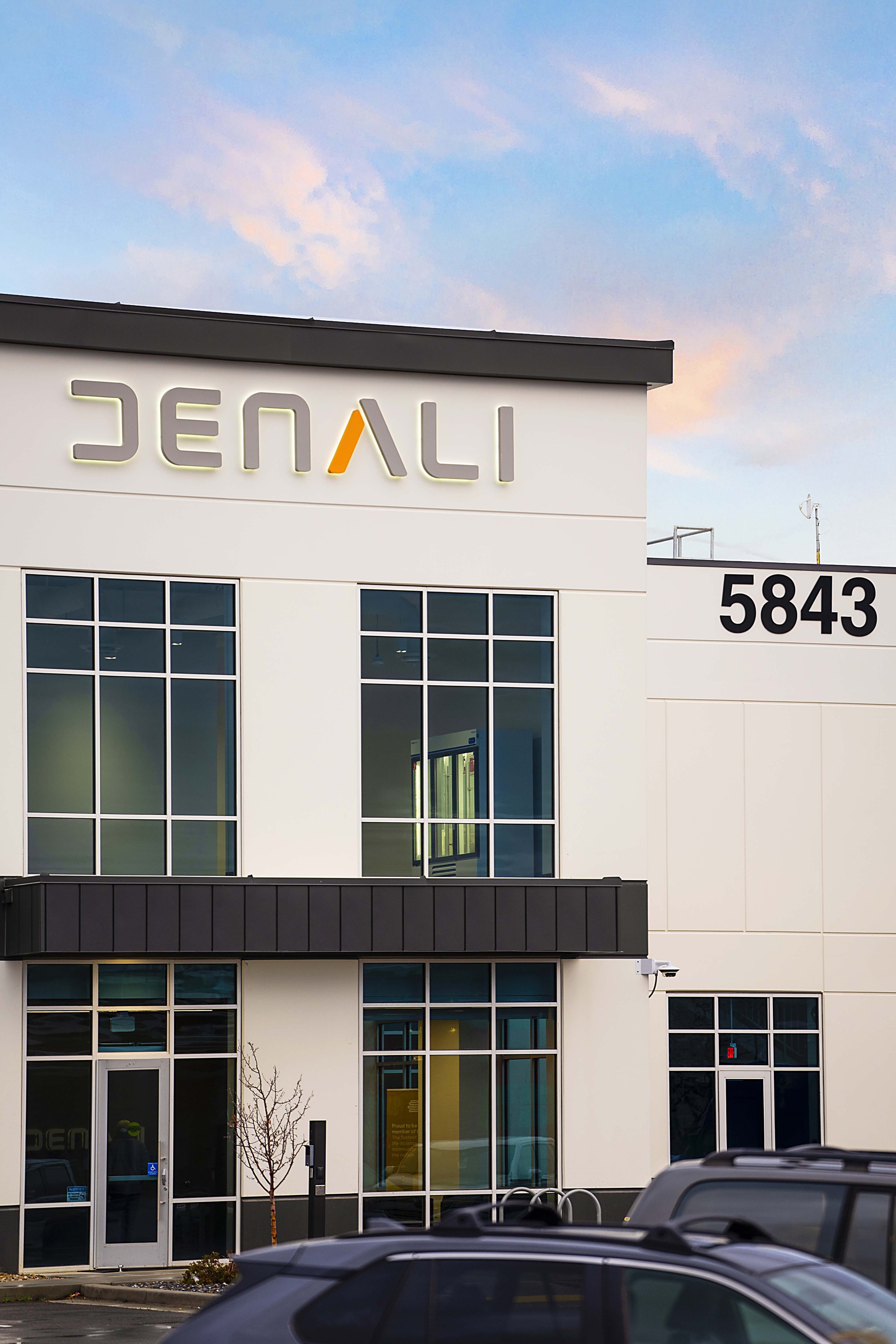 Front view of a modern commercial building with the sign 'DENALI' and the number 5843, with parked cars in the foreground and a partly cloudy sky in the background.