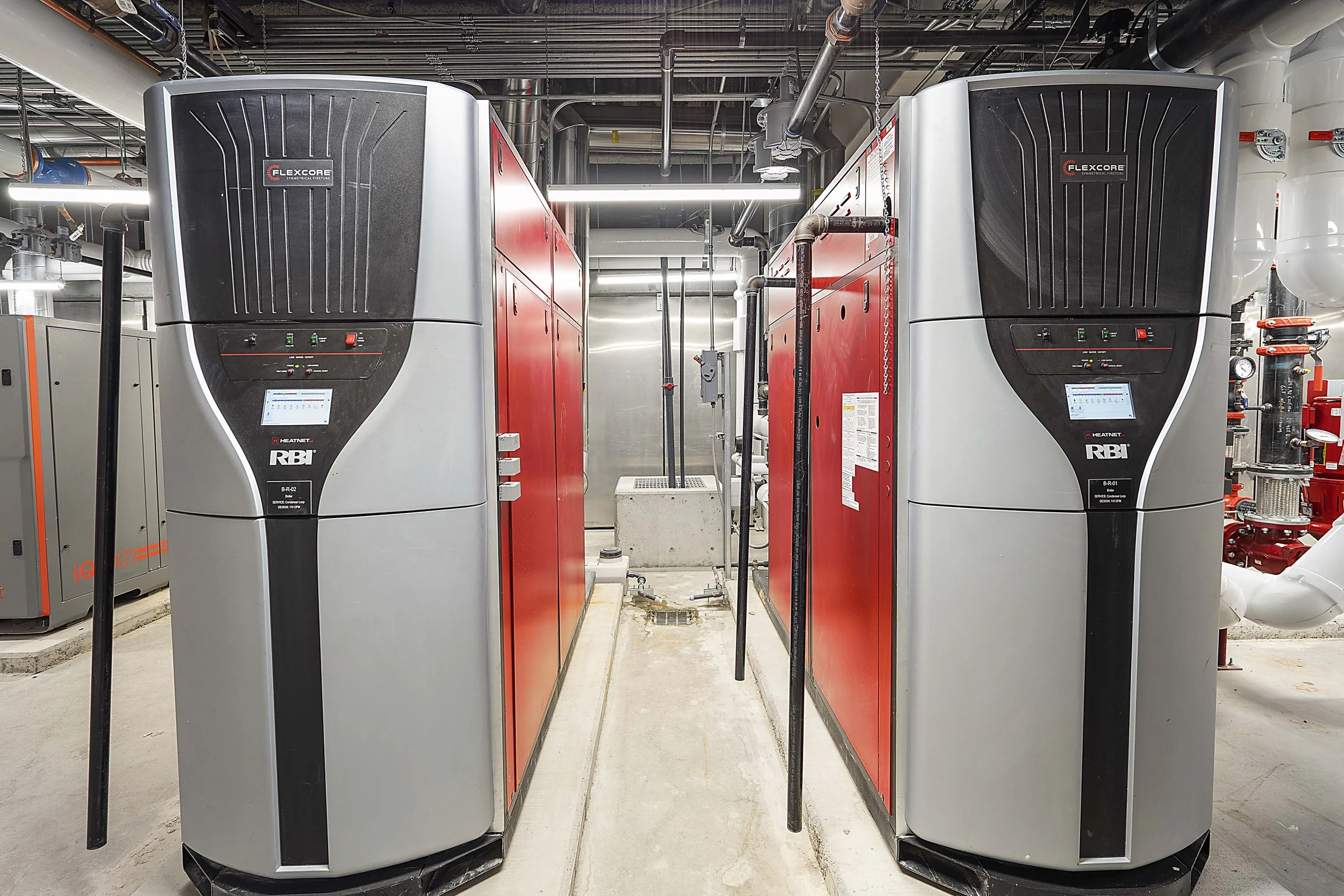 Two large industrial boilers with black and gray covers, red panels, and touchscreen displays, situated in an industrial utility room.