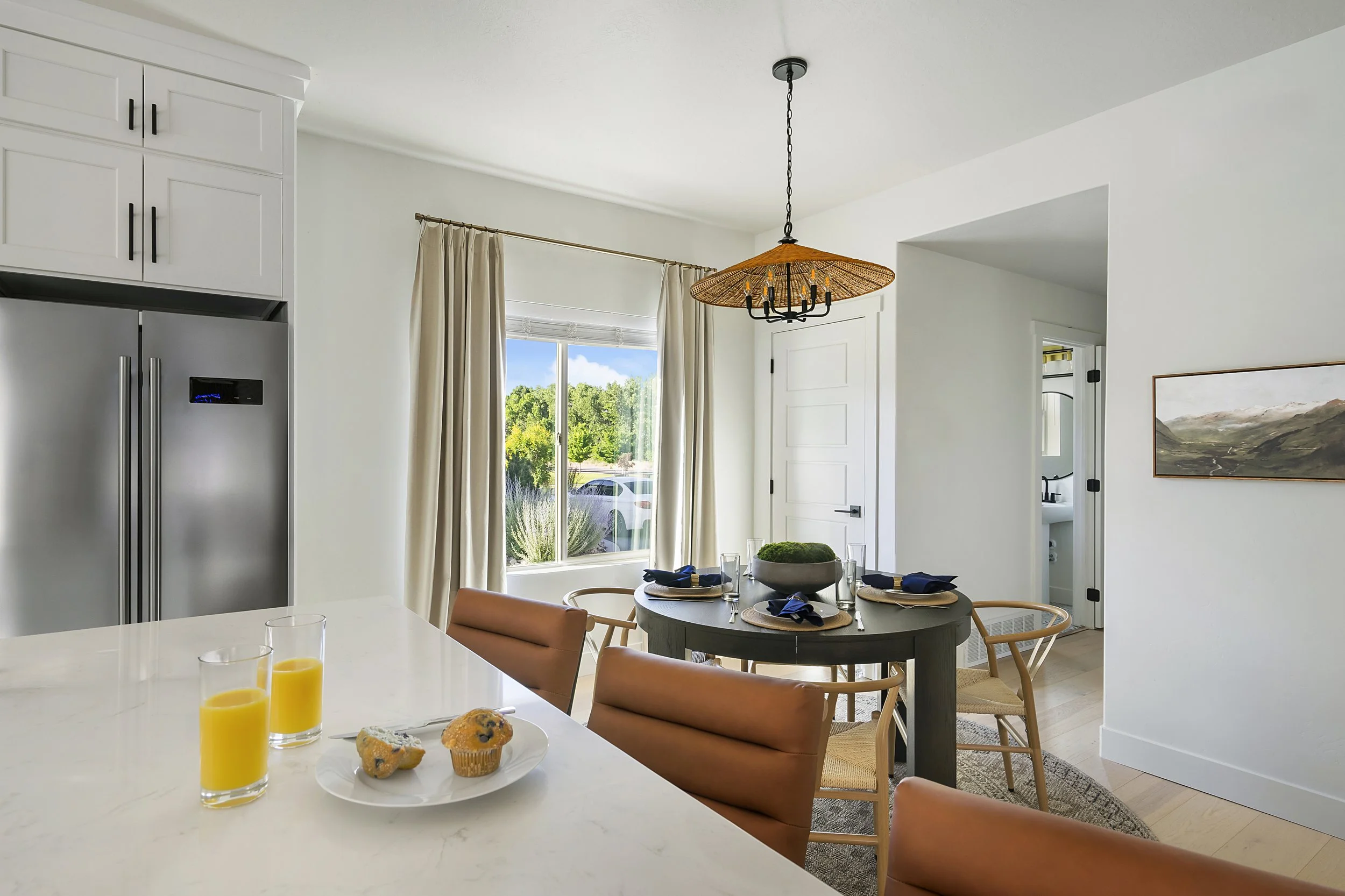 Bright dining area with a round table set for four, surrounded by chairs, in front of a window with beige curtains showing greenery outside. A modern chandelier hangs above the table. In the foreground, a white countertop has two glasses of orange ju