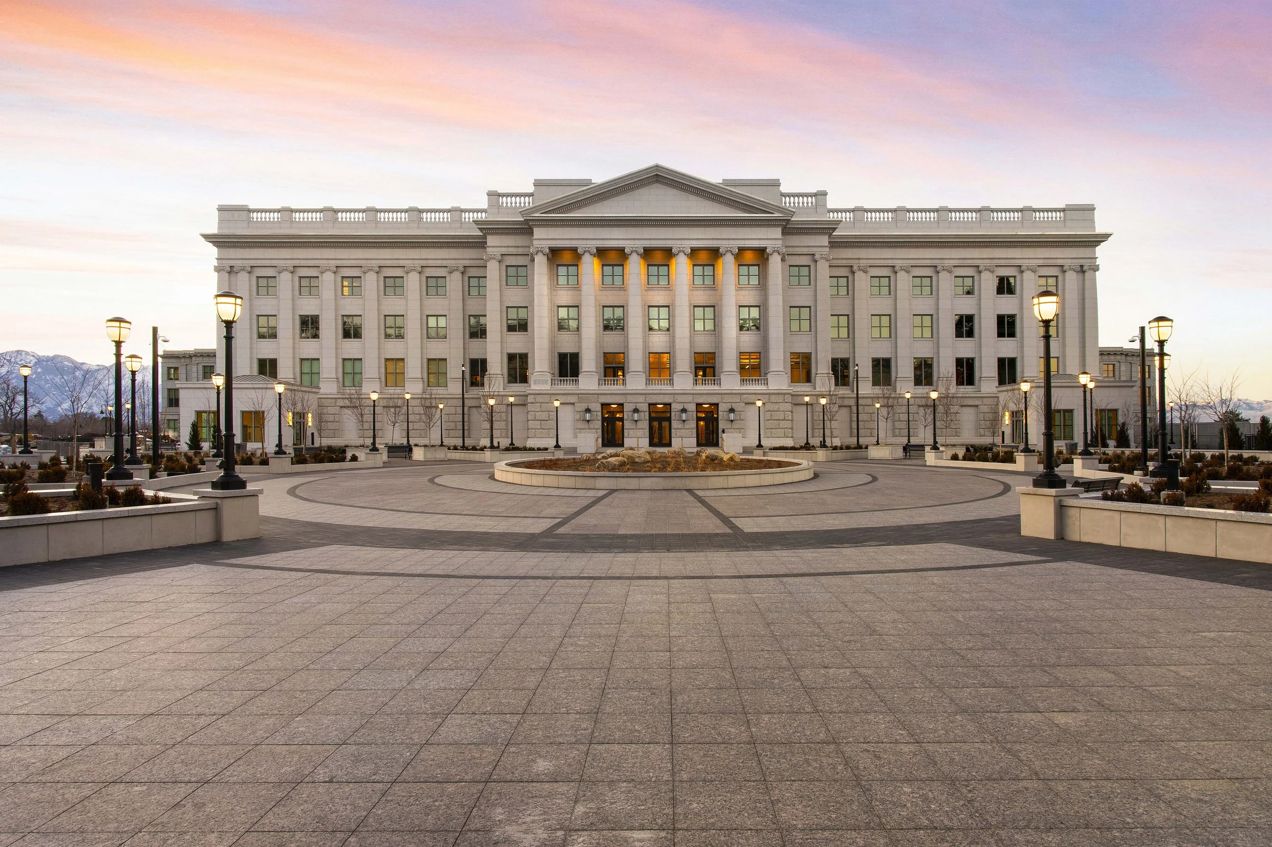 A large, white government or institutional building with classical architectural features, illuminated by street lamps, under a pink and purple sunset sky.